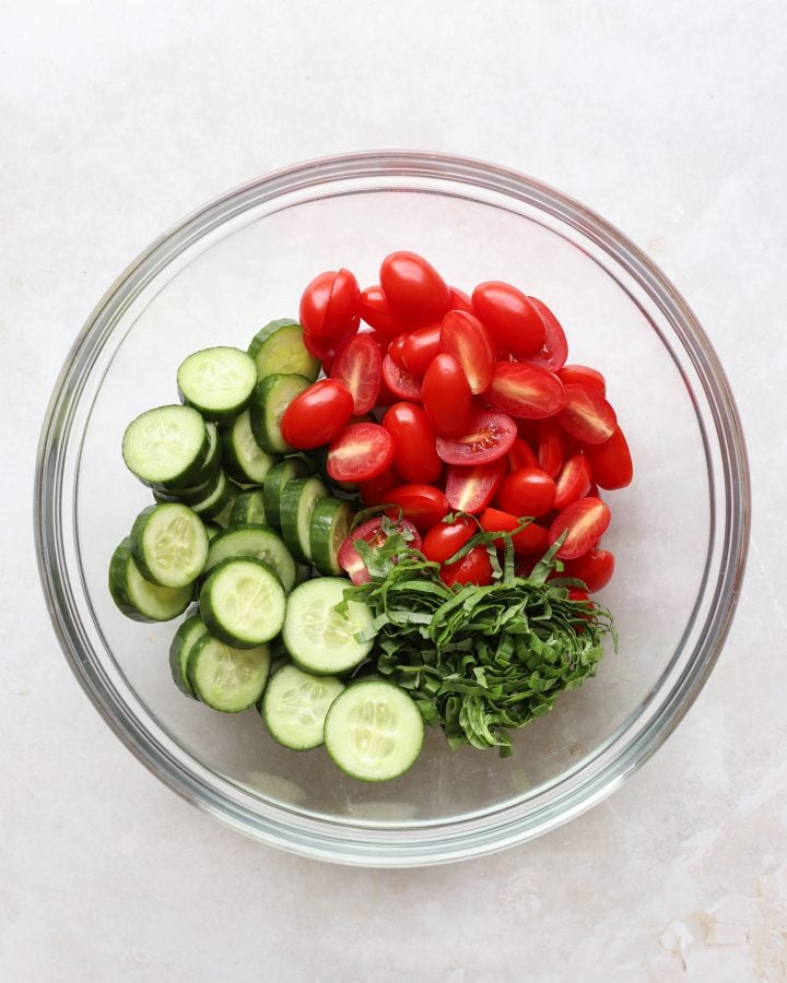Sliced tomatoes, sliced cucumbers, fresh thinly sliced basil in a glass bowl. 