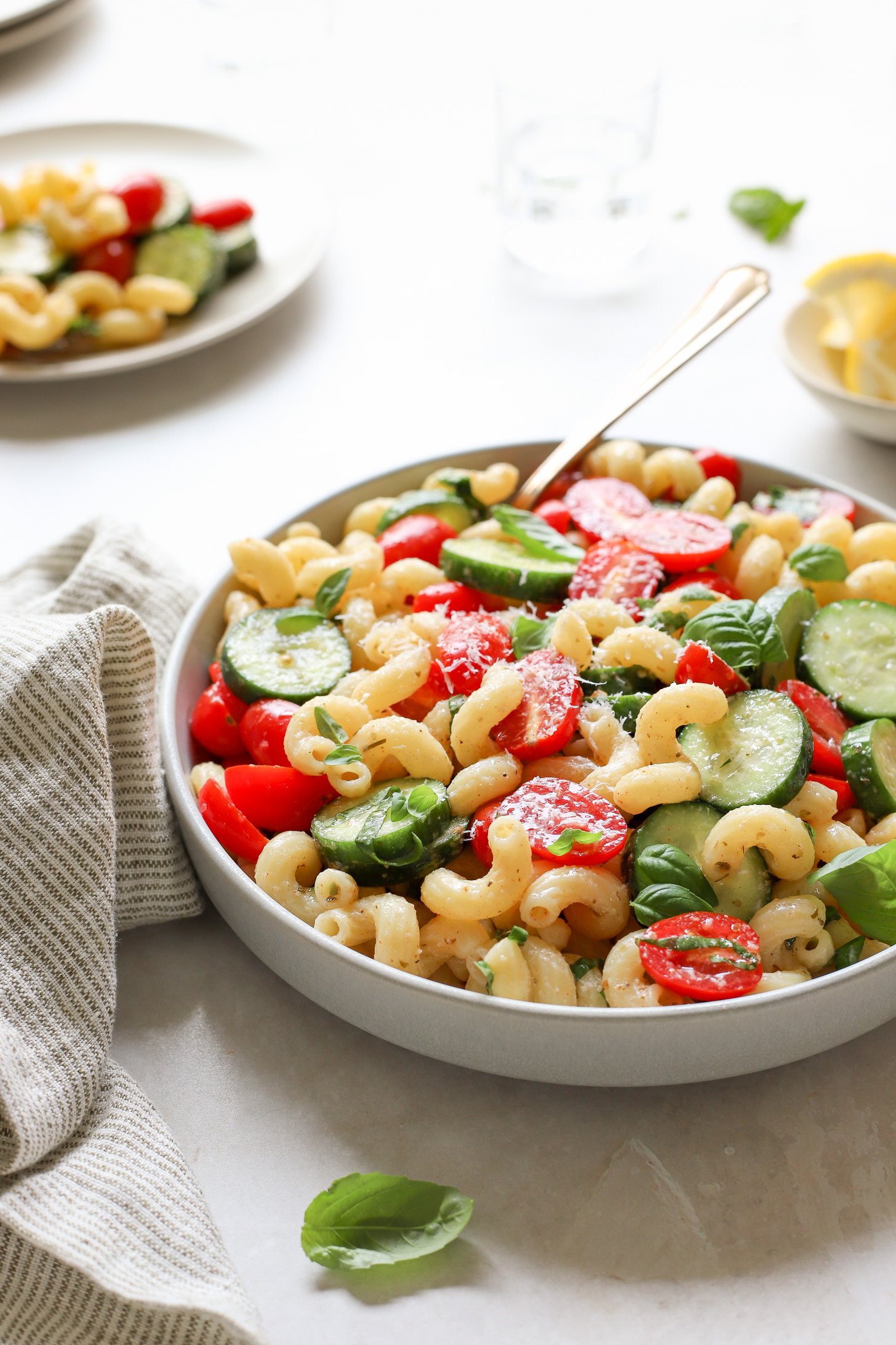 A bowl of cucumber tomato pasta salad in a white bowl on a table with a striped napkin. There is a spoon in the bowl and there is a serving of salad on a plate in the corner,  bowl of sliced lemons, plates and a glass of water on the table next to the bowl. 
