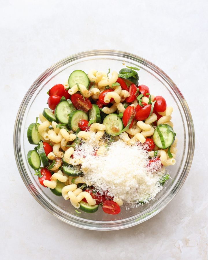 Cucumber tomato pasta salad in a bowl with grated parmesan cheese on top before being mixed together. 