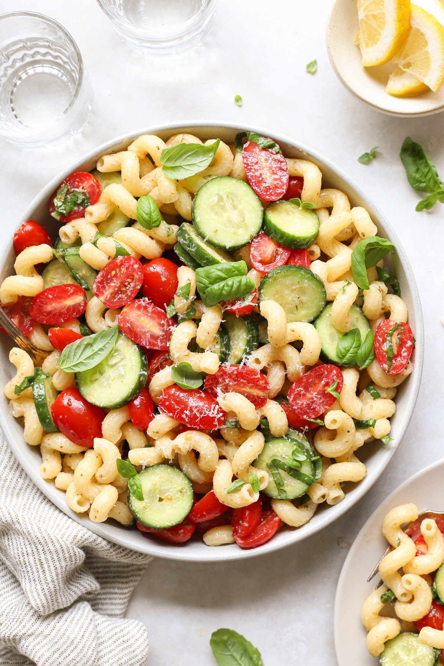 Cucumber Tomato pasta salad in a white bowl on a white table with two glasses of water, a bowl of sliced lemons and a striped napkin. There is a plate with a serving of pasta salad in the corner of the picture.