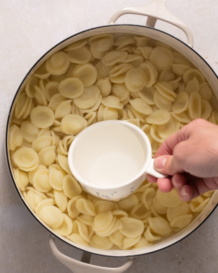 Pasta cooking in a pot. A hand is using a measuring cup to scoop some pasta water out.