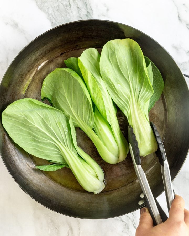 Halved bok choy in an oiled skillet, cut-side down. A person is using tongs to place one of the bok choy halves in the pan.