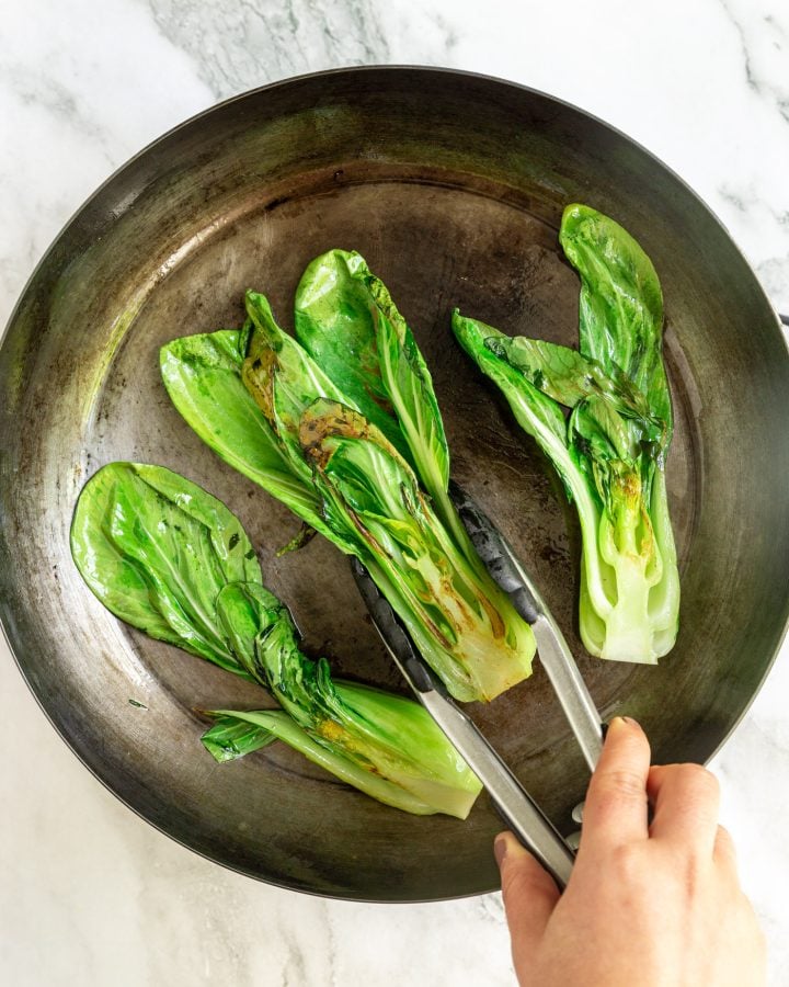 Halved seared bok choy in a skillet. A person is using tongs to flip over the middle piece.