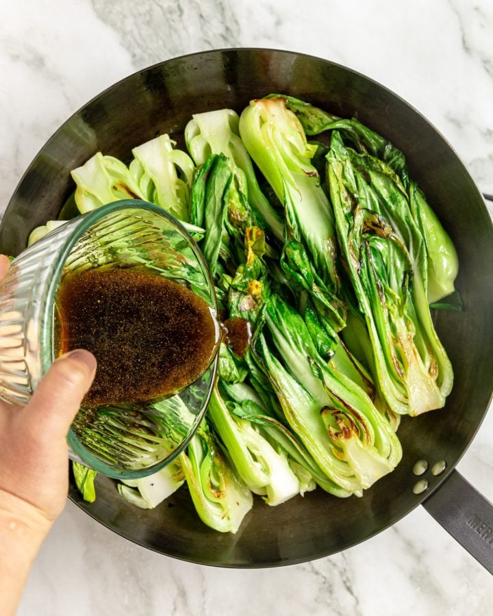 A person pouring sesame vinaigrette over seared bok choy in a skillet.