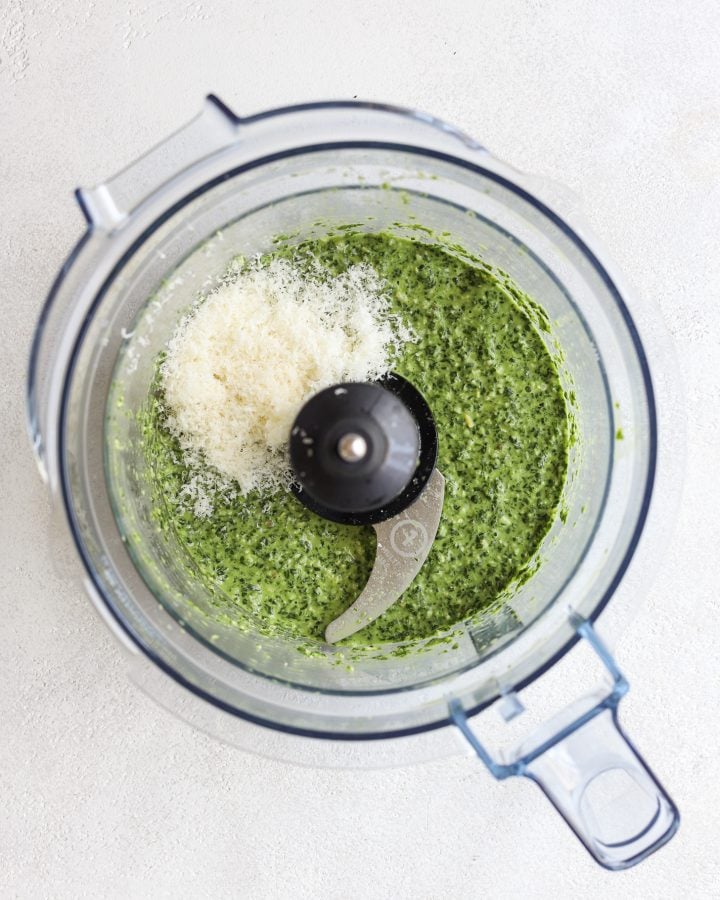 Pureed spinach pesto in a food processor with grated parmesan cheese on top before being mixed in.