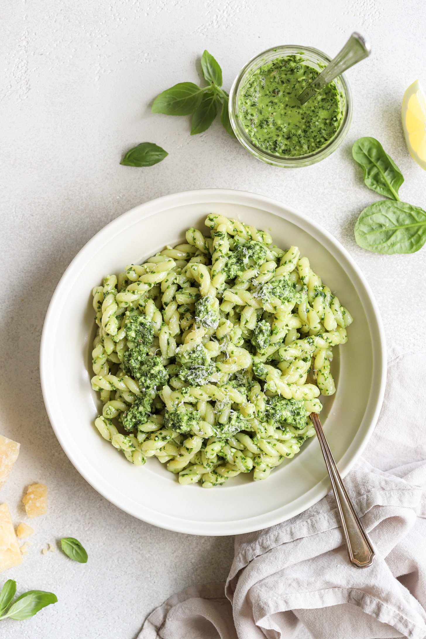 Spinach pesto tossed with cooked pasta in a white bowl with a fork. There is a white napkin, the jar with pesto, and ingredients from the pesto scattered around the bowl on the table.