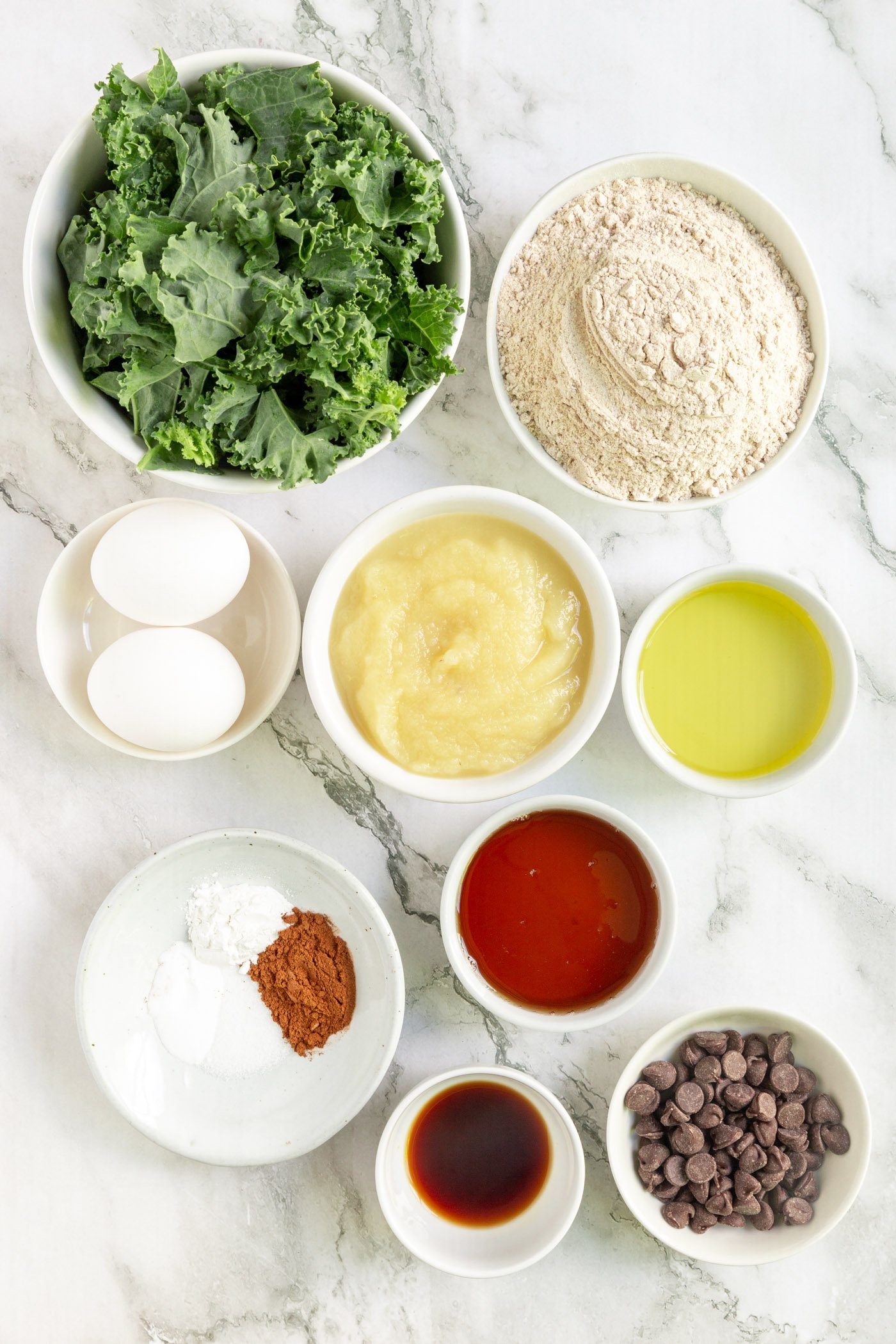 Ingredients for sweet kale muffins in bowls on a white marble table. 