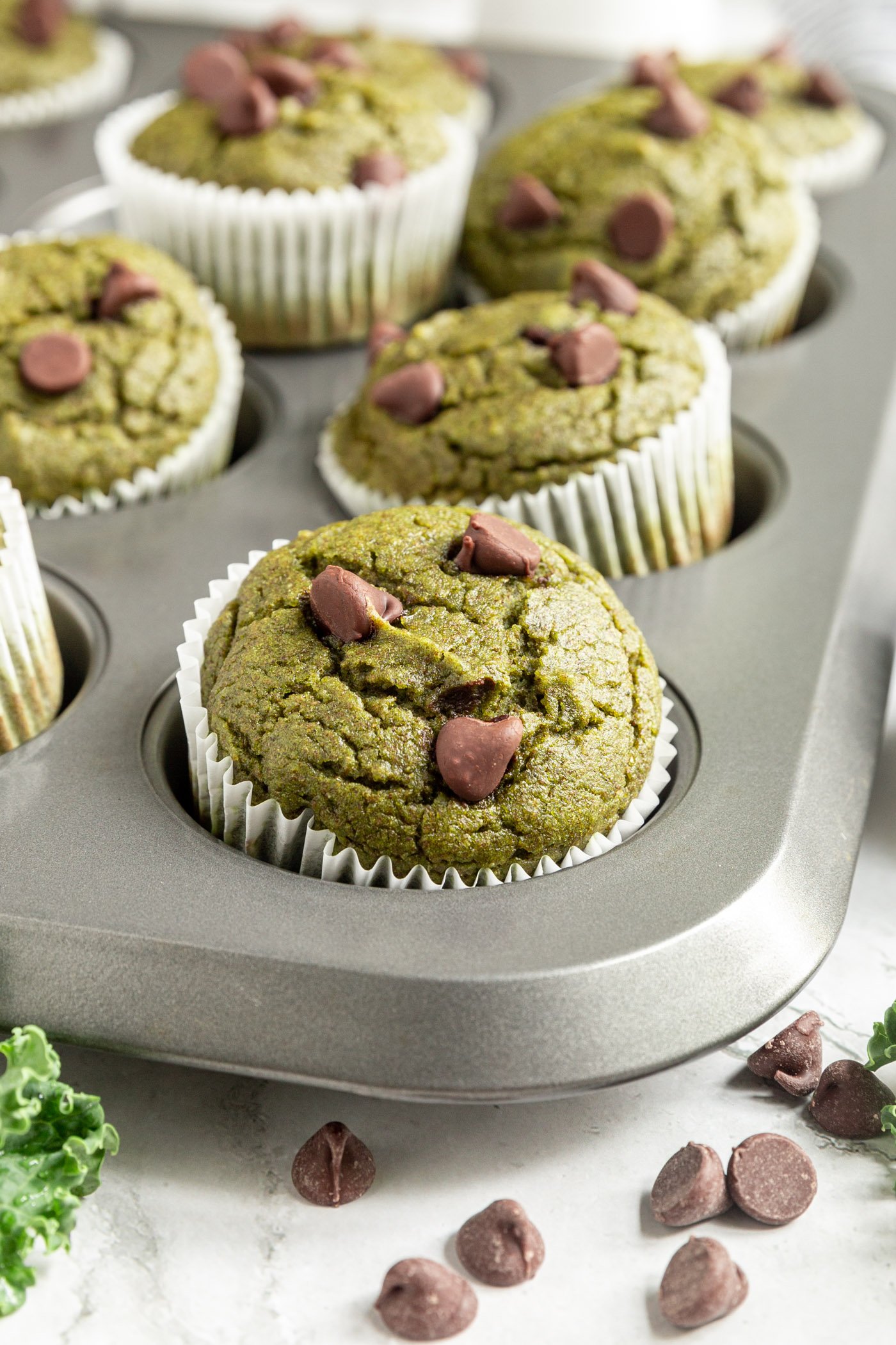 Baked kale muffins in a muffin tin on a table. There are chocolate chips scattered around the tin on the table.