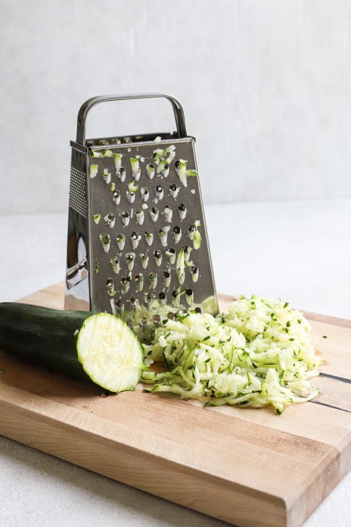 A zucchini half grated on a cutting board with a box grated and pile of grated zucchini. 