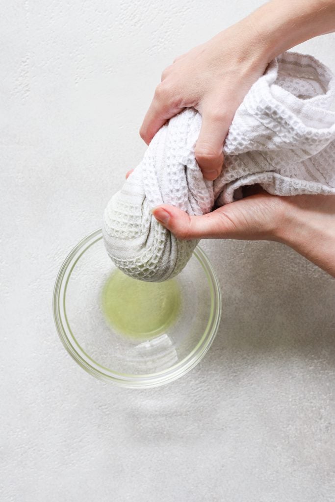 A person squeezing out the grated zucchini  in the towel over a glass bowl.