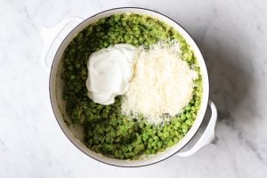 Cooked and mashed peas in a white pot. There is grated parmesan cheese and Greek yogurt on top.