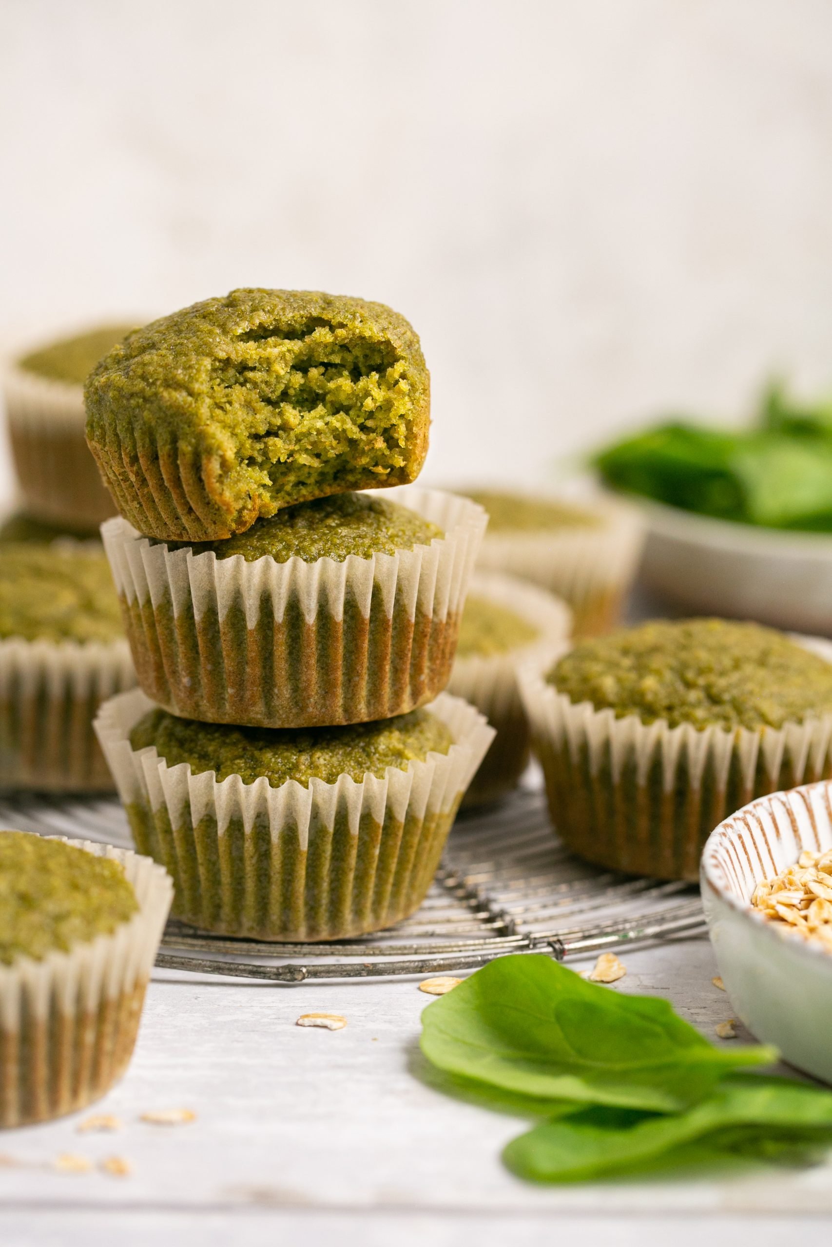 3 green smoothie muffins stacked on top of one another. The top one has a bite out of it. You can see more muffins and muffin ingredients blurred in the background. 