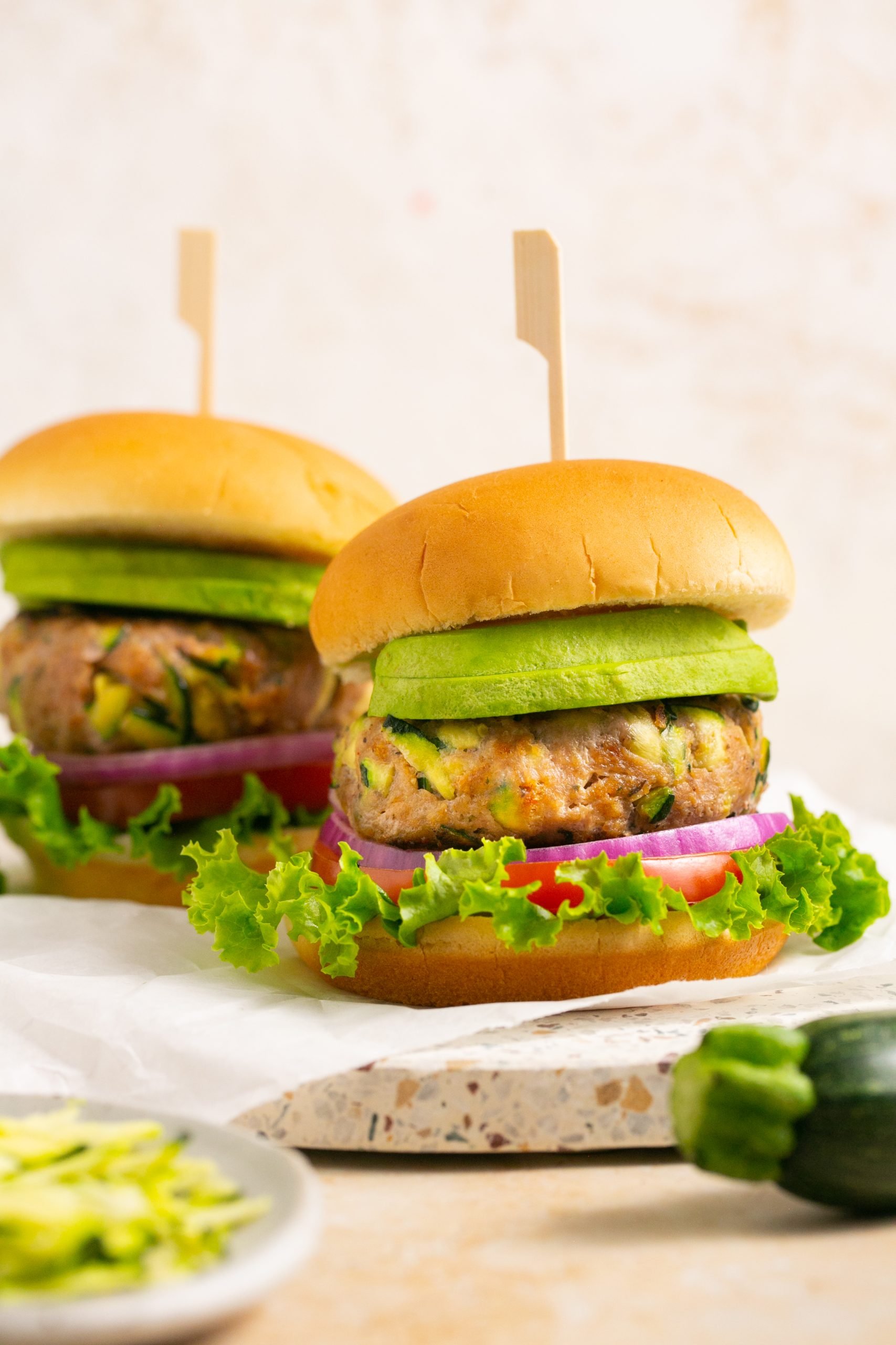 Two zucchini turkey burgers on buns with lettuce, tomato, red onion and avocado