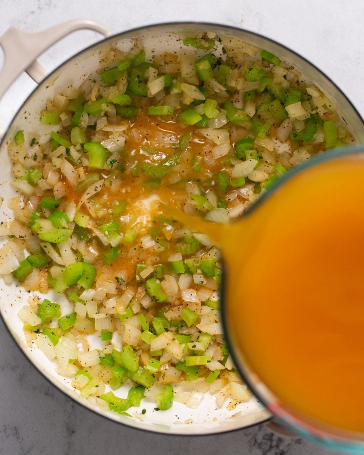 Vegetable broth being poured into a Dutch oven with sauteed and seasoned celery and onions. 