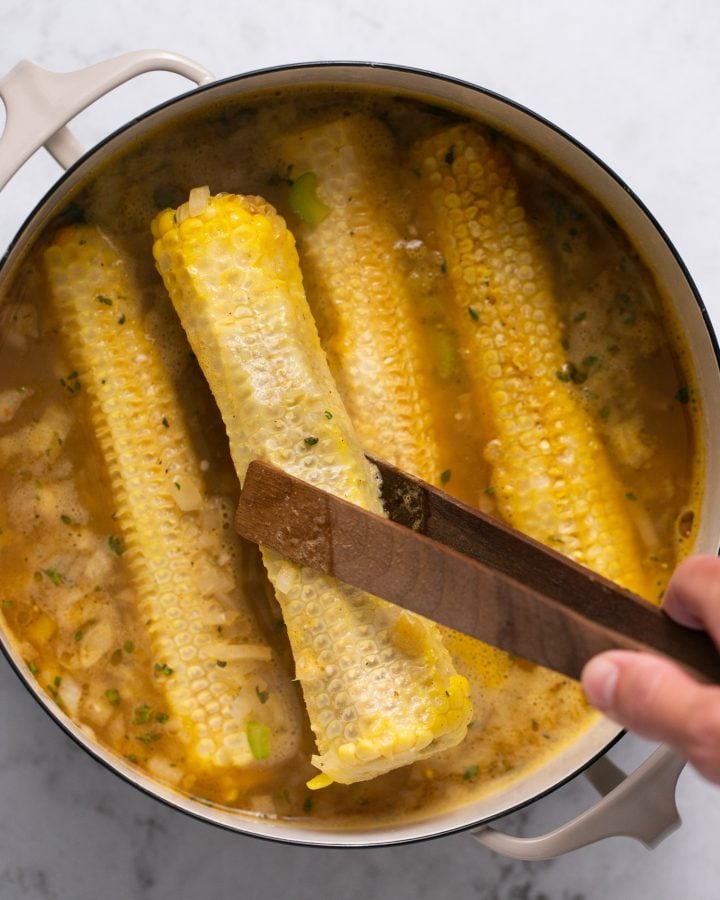 Tongs being used to remove corn cobs from a Dutch oven filled with broth and sauteed veggies. 