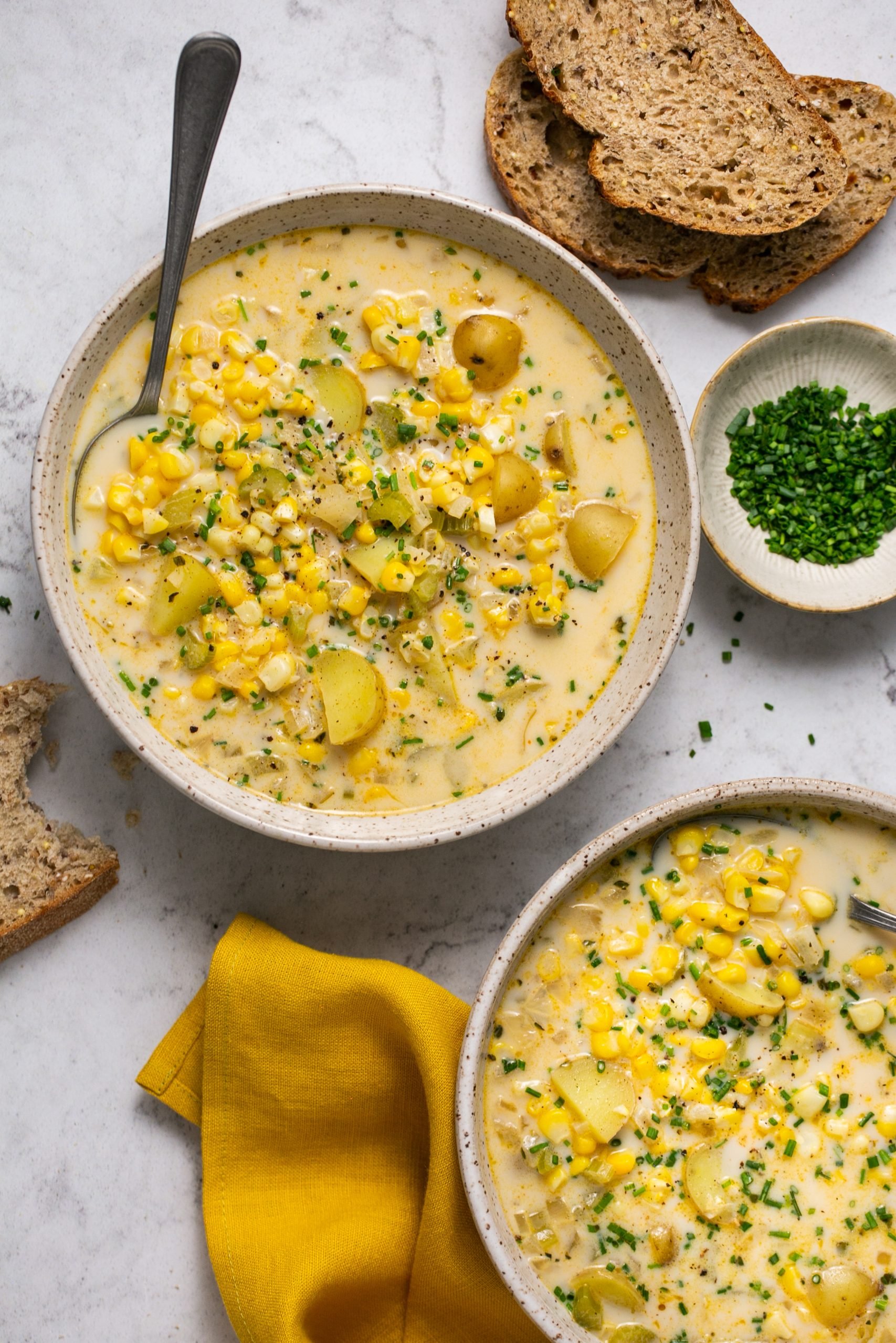 A bowl of corn and potato chowder with a spoon. There are slices of bread, a yellow napkin and a bowl of chives next to the bowl on the table. 