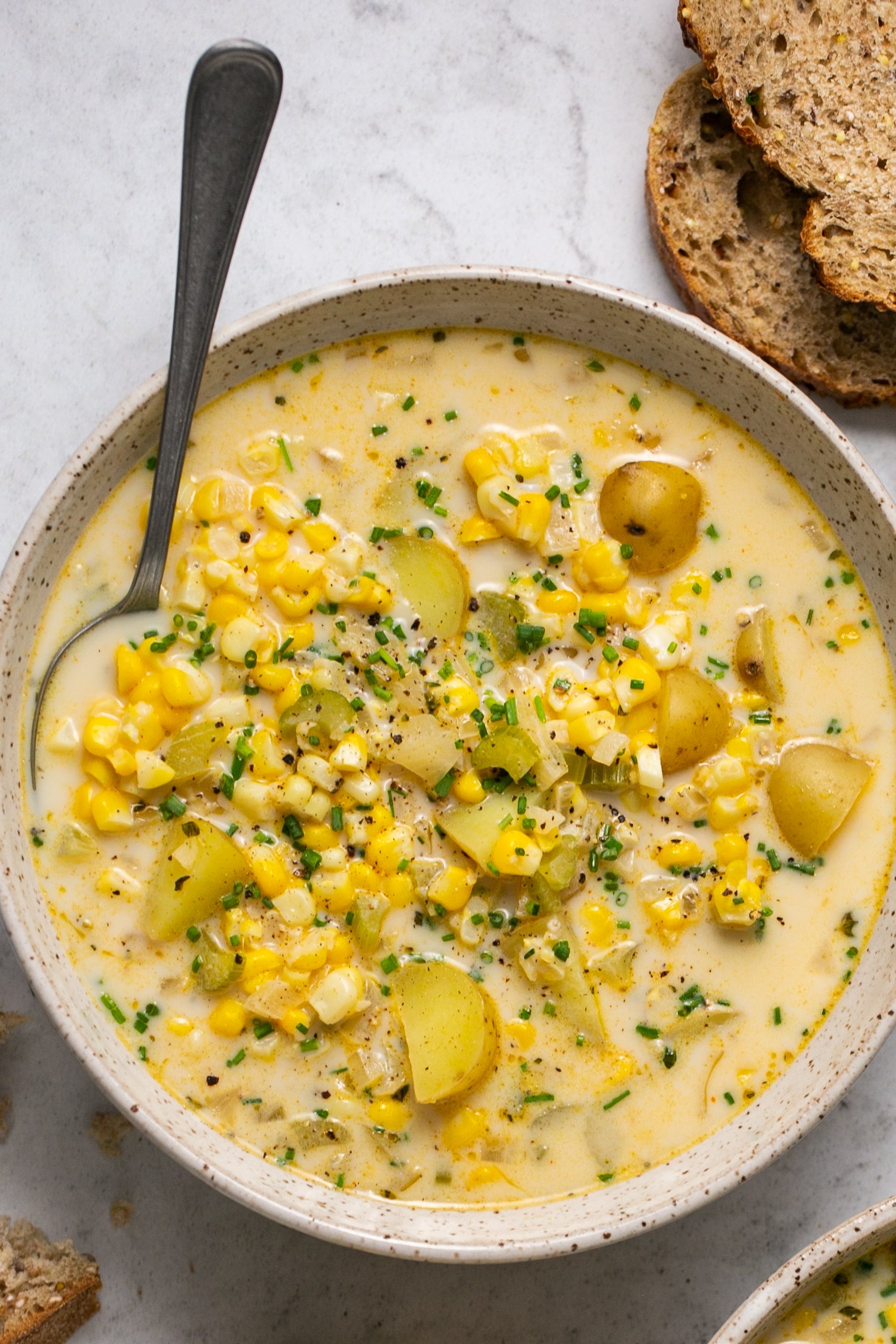 A bowl of corn and potato chowder with a spoon. There are slices of bread next to the bowl.