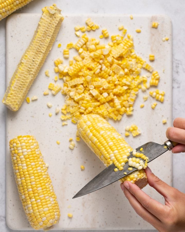 A person using a large knife to cur corn off of the cob on a cutting board. 