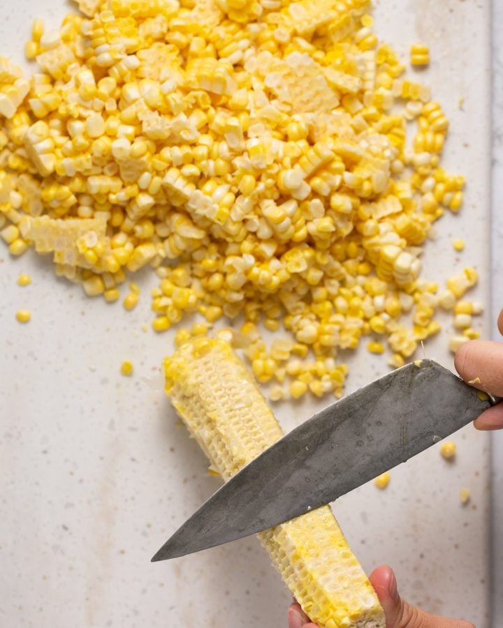 A person using the back of a large knife to get the corn juice out fo the cob.