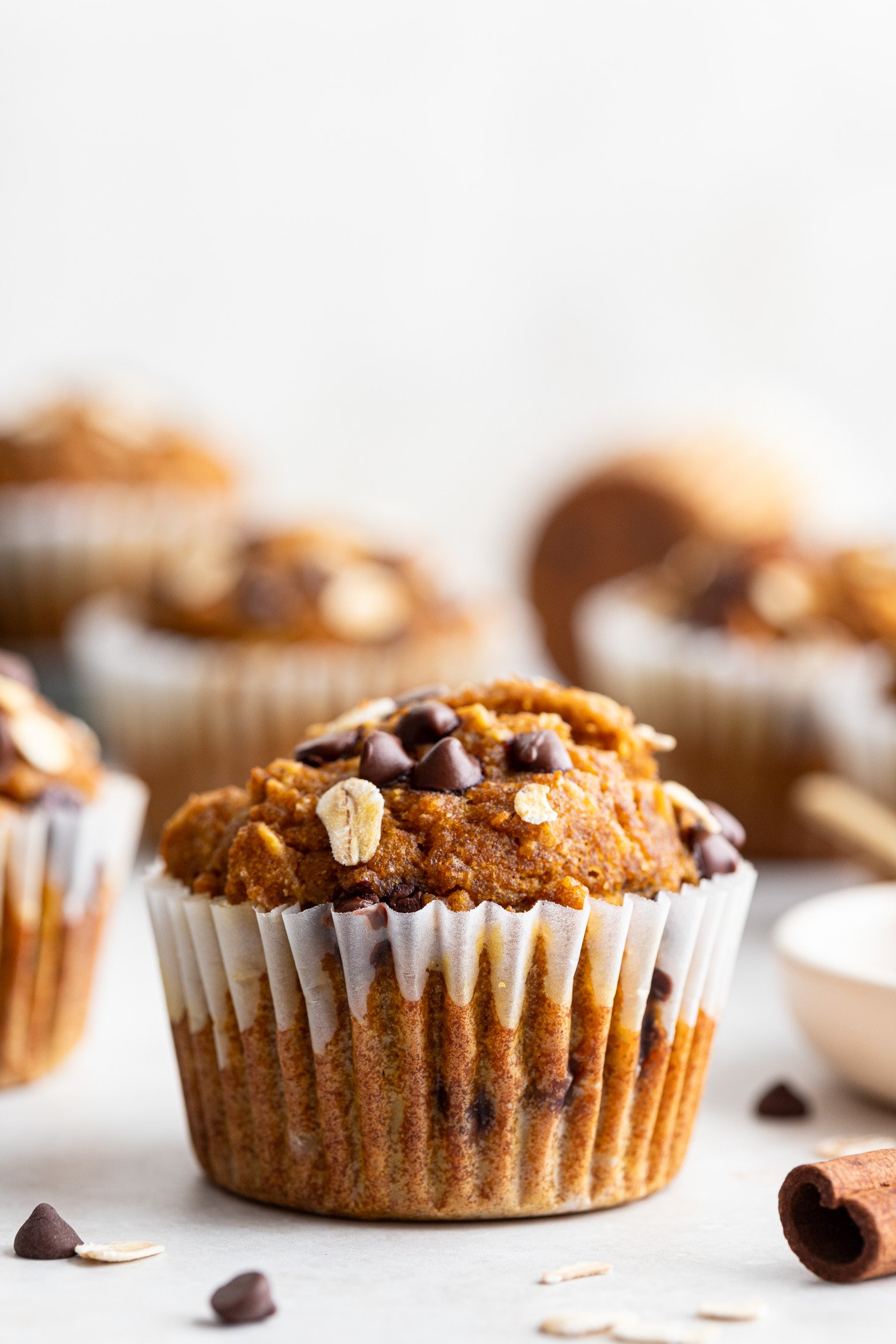 Muffin in a muffin liner with chocolate chips and oatmeal on top sitting on a white surface. More muffins are out-of-focus in the background. A few chocolate chips, oats, and a cinnamon stick are strewn on the counter.
