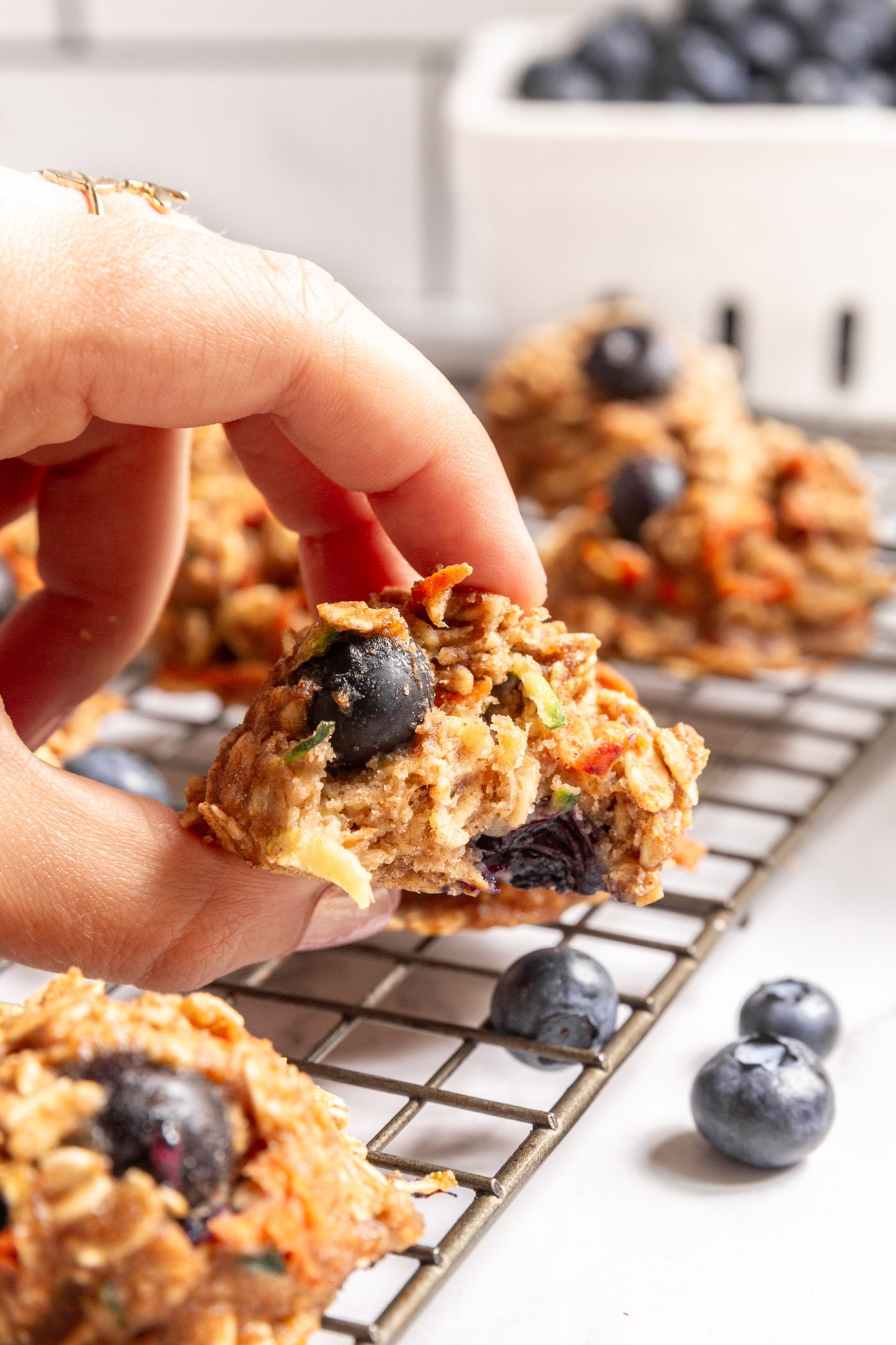 A hand holding a veggie-loaded breakfast cookie. It has a bite taken out of it and there are more cookies blurred on the baking sheet behind it. 
