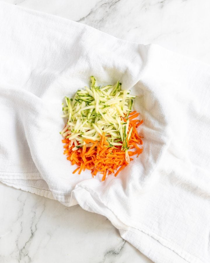 Grated zucchini and gated carrots in the middle of a white towel.