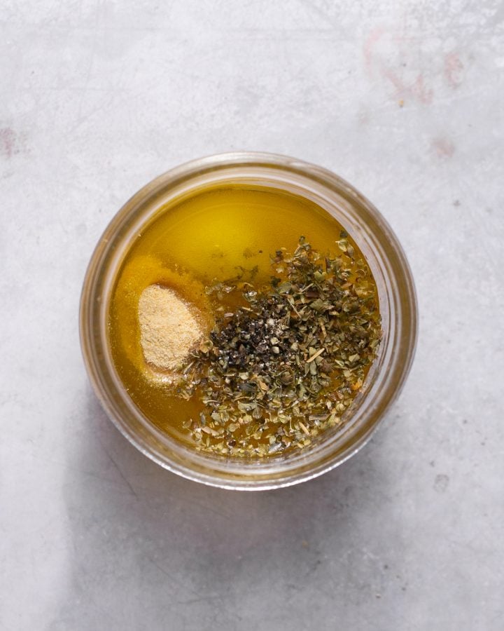 Spices (garlic powder, oregano, salt, and pepper) sitting on top of the vinegar, honey, and olive oil in a small glass jar.