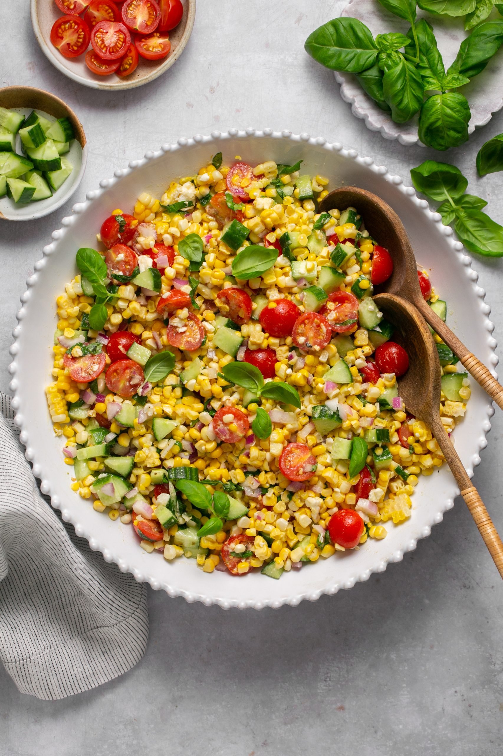 Corn salad in a white serving bowl. 2 wooden serving spoon are sitting in the corn salad. Basil sprigs, sliced tomatoes, and chopped cucumbers are set to the side in individual bowls.