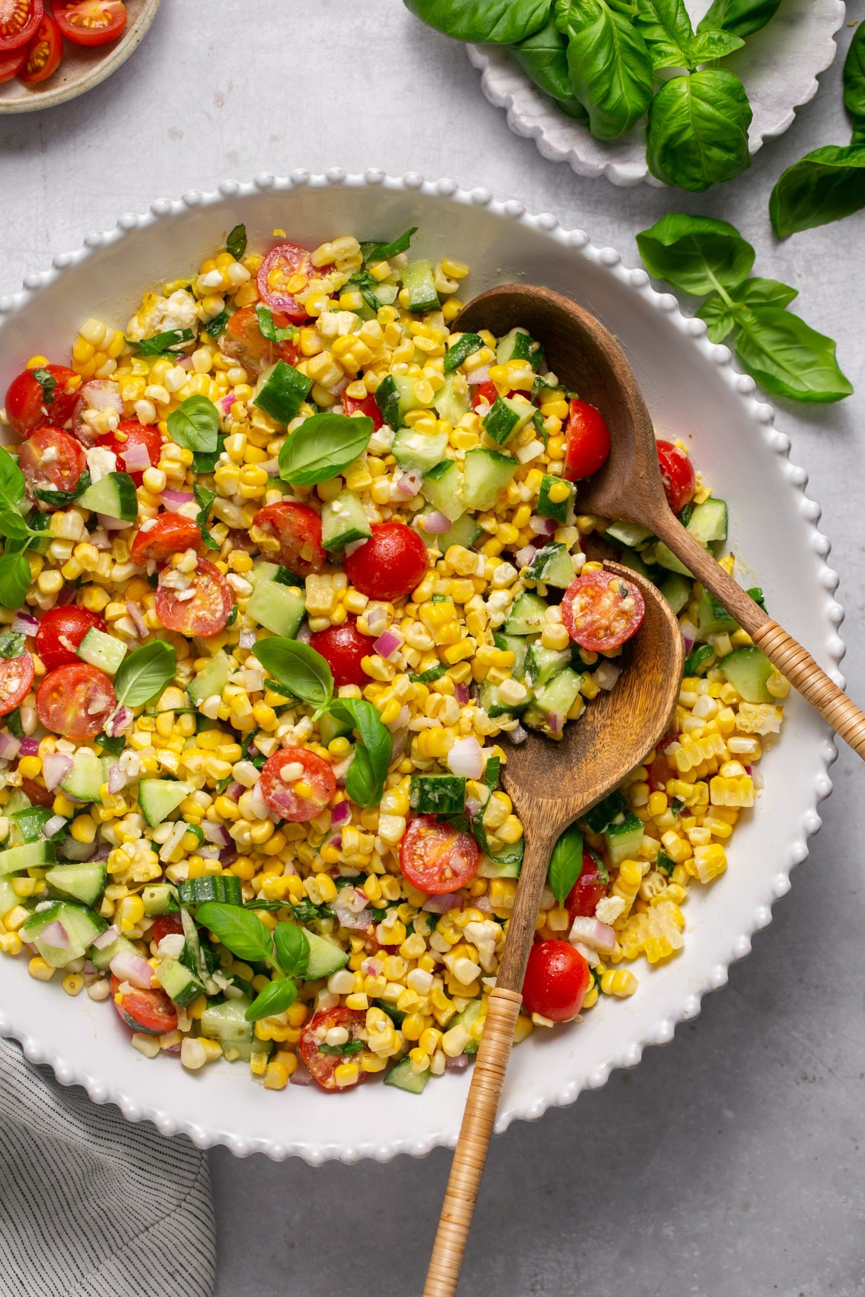 Corn salad in a white serving bowl. 2 wooden serving spoon are sitting in the corn salad. Basil sprigs and sliced tomatoes are set to the side in individual bowls.
