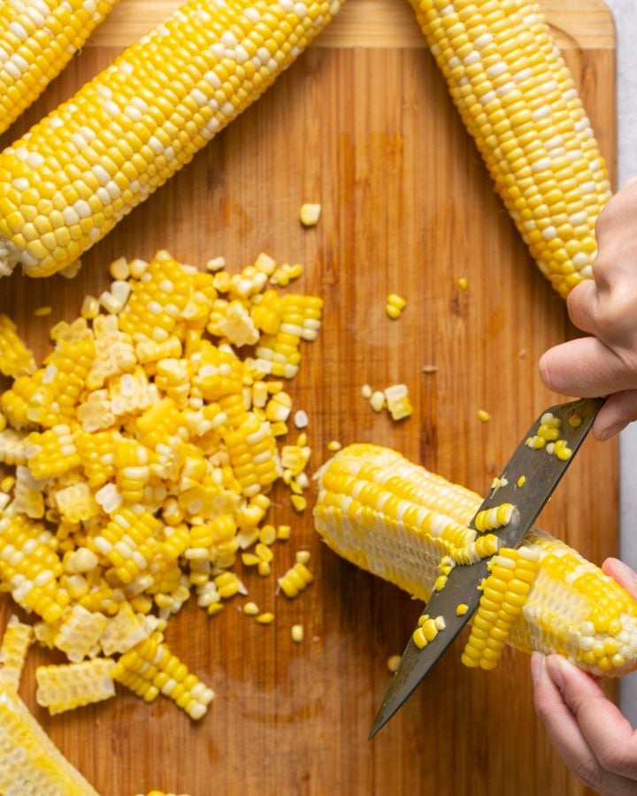 Hand with knife cutting corn kernels off of a cooked ear of corn onto a wooden cutting board. Three additional ears of corn are set to the side.