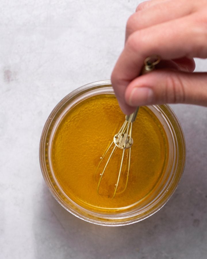 Hand holding a whisk, whisking together vinegar, honey, and olive oil in a small glass container.