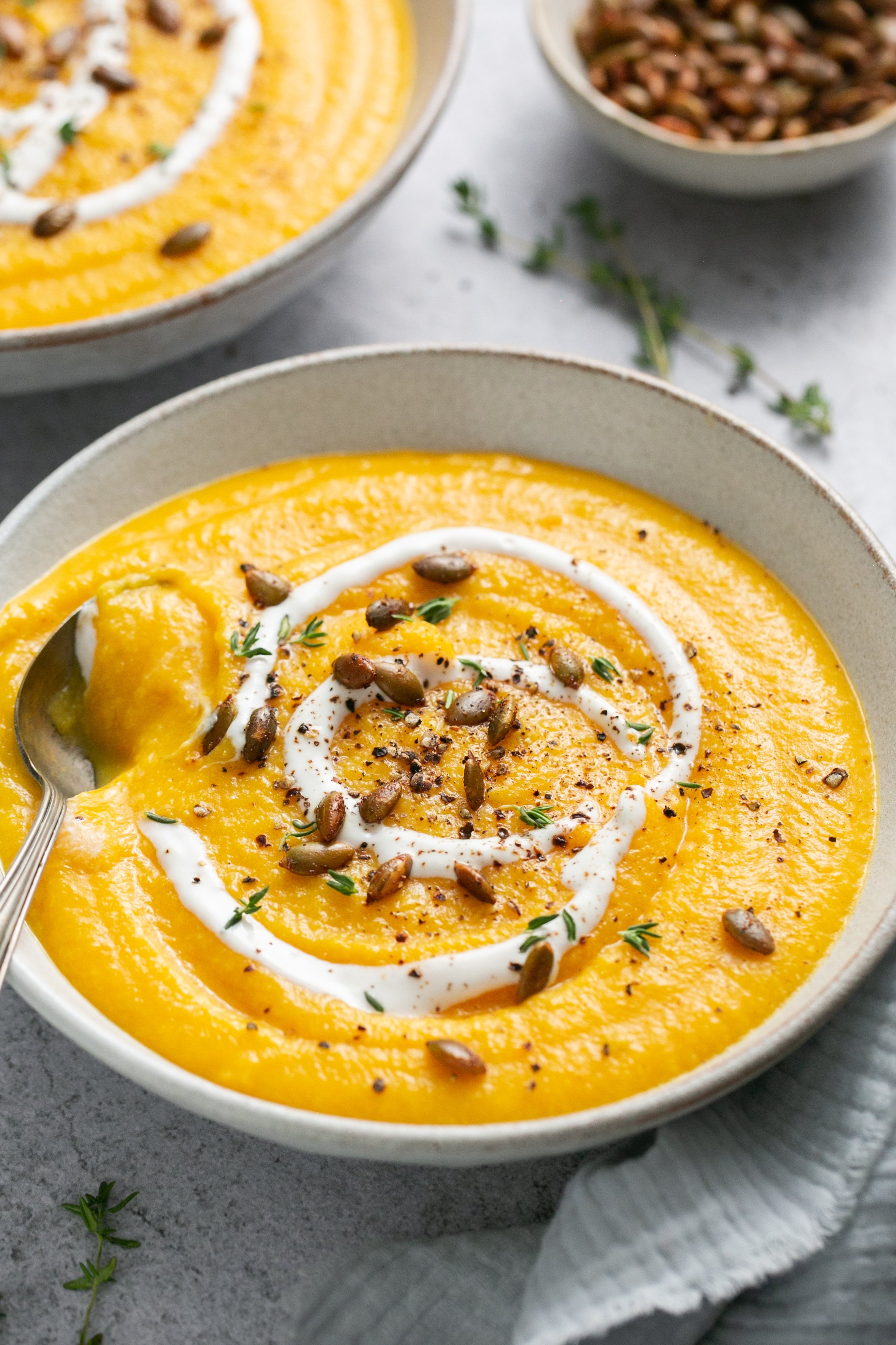 A bowl of creamy butternut squash soup on a table with a spoon in the bowl. It is garnished with pumpkin seeds, pepper, extra yogurt and thyme. 