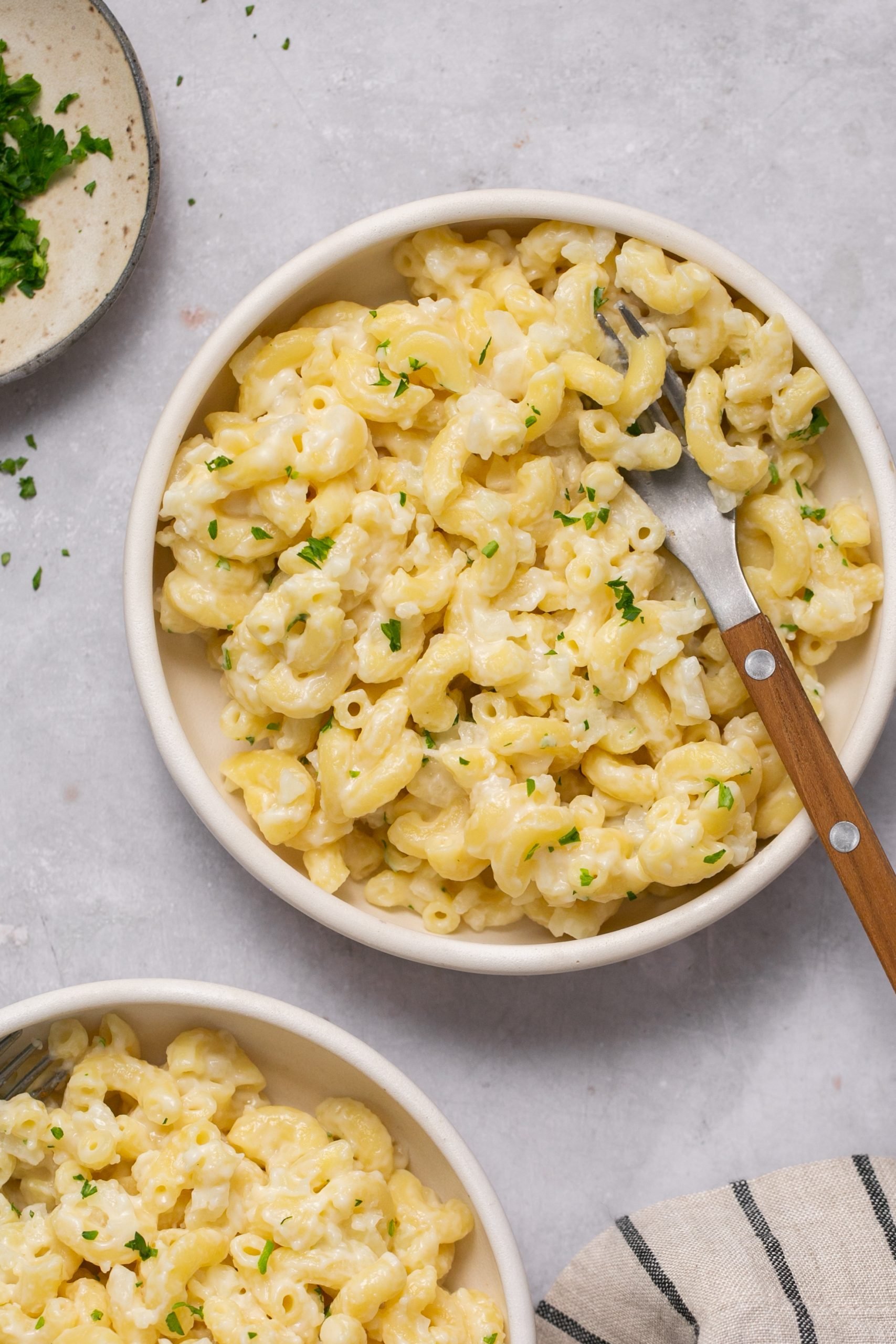 Two bowls of cauliflower mac and cheese on a table with a little chopped parsley on top. One bowl has a fork in it. 