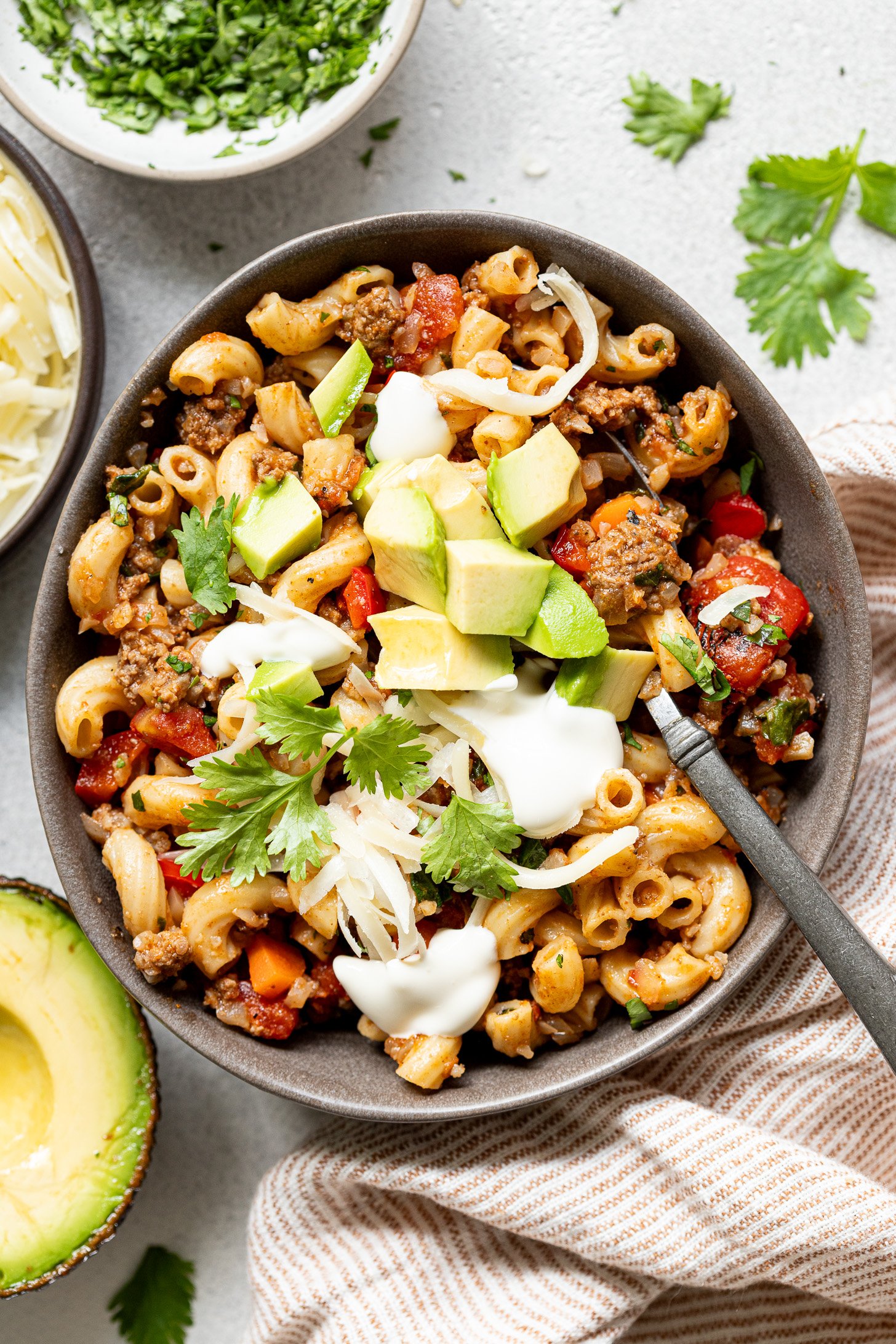 A bowl of veggie-loaded taco pasta in a gray bowl with sour cream, avocado, grated cheese, and fresh cilantro on top. There is a fork in the bowl. There is a striped napkin next to the bowl and bowls with toppings on the table.