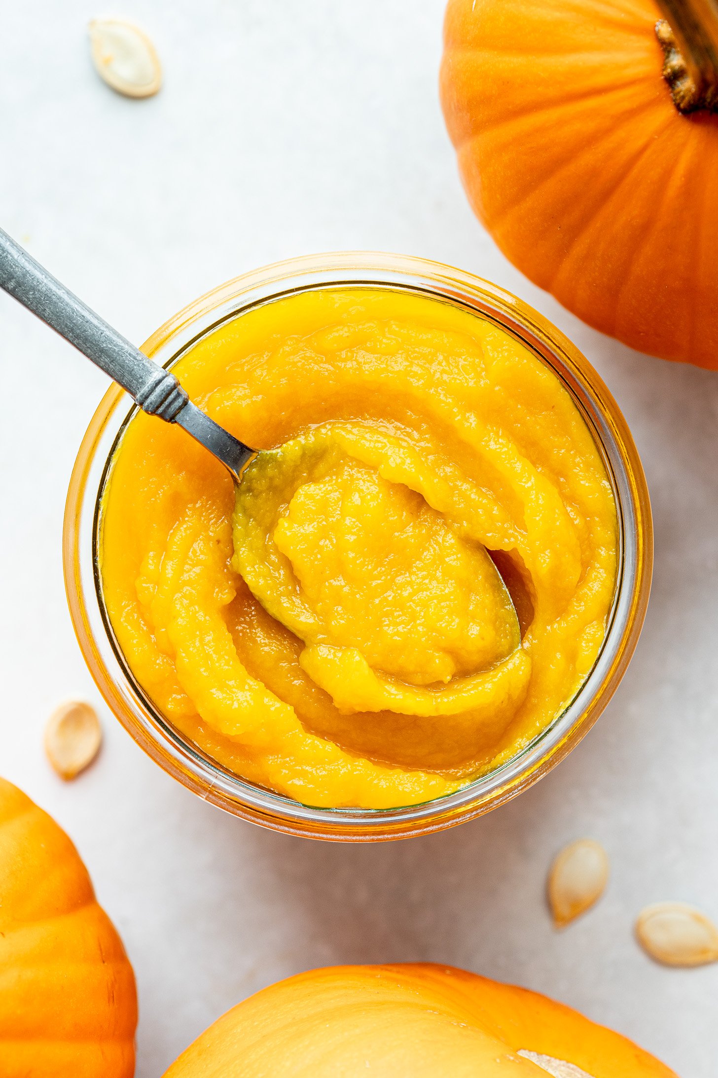 A spoon sitting in a glass jar of pumpkin puree sitting on a white countertop. Pumpkin seeds strewn about on countertop. Three pumpkins set to the side.