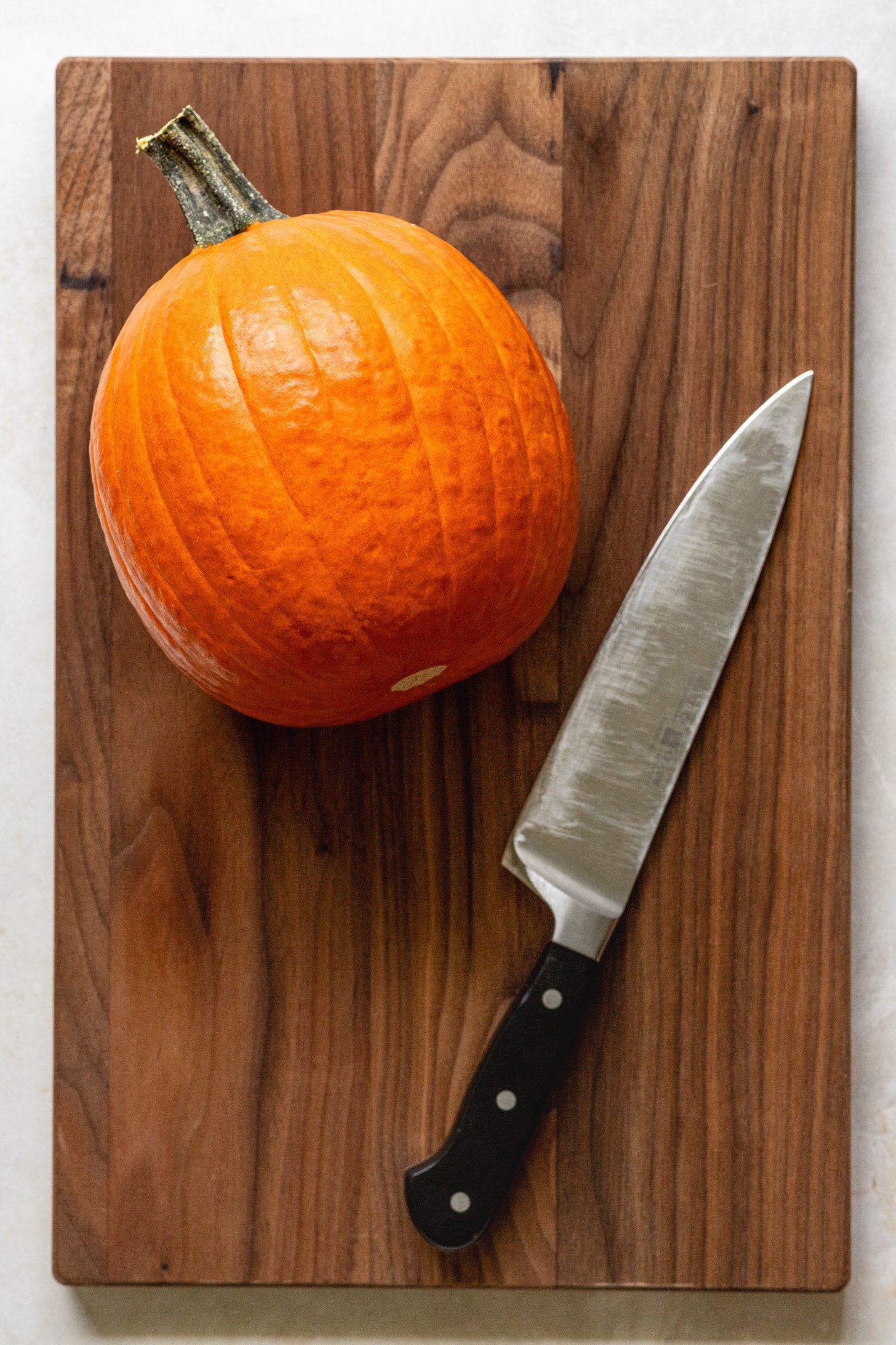 A small pumpkin and chef's knife sitting atop a wooden cutting board.