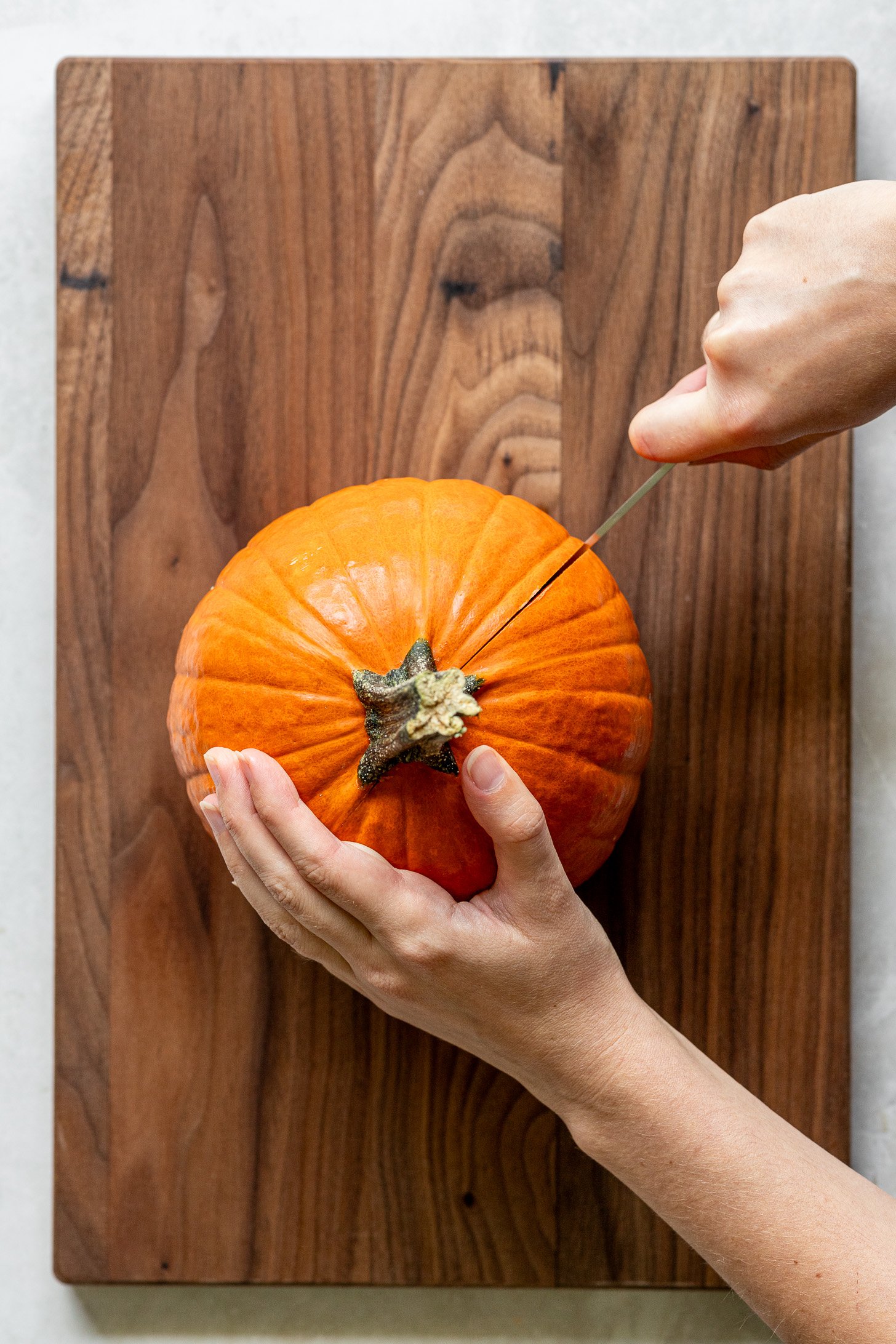 Pair of hands holding onto a small pumpkin and slicing into the pumpkin that sits on top of a wooden cutting board. 