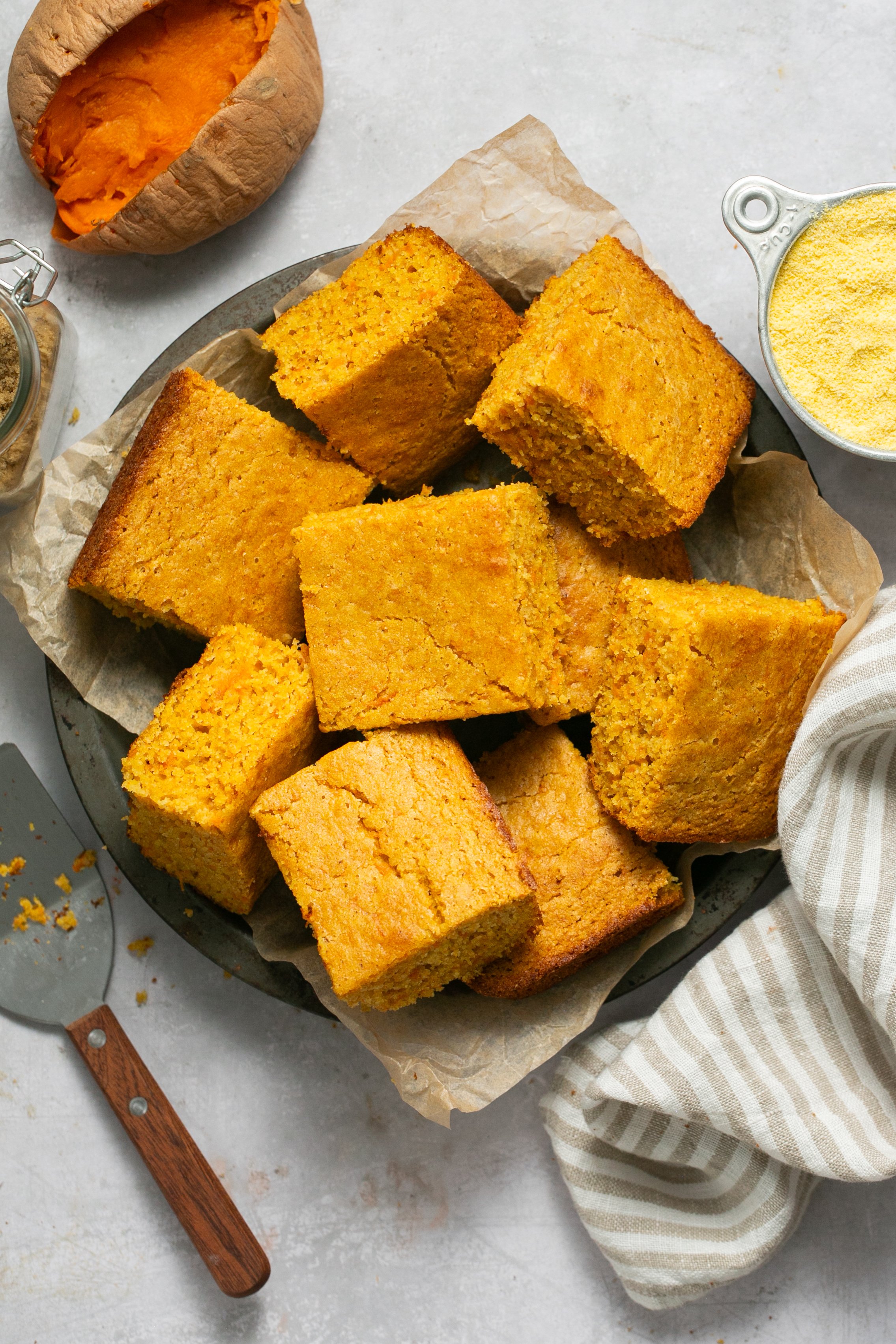 Cut pieces of sweet potato cornbread in a baking dish lined with parchment paper on a white table. There is a measuring cup filled with cornmeal, a cooked sweet potato, a beige and white linen napkin, and a serving spatula set off to the side on the table as well.