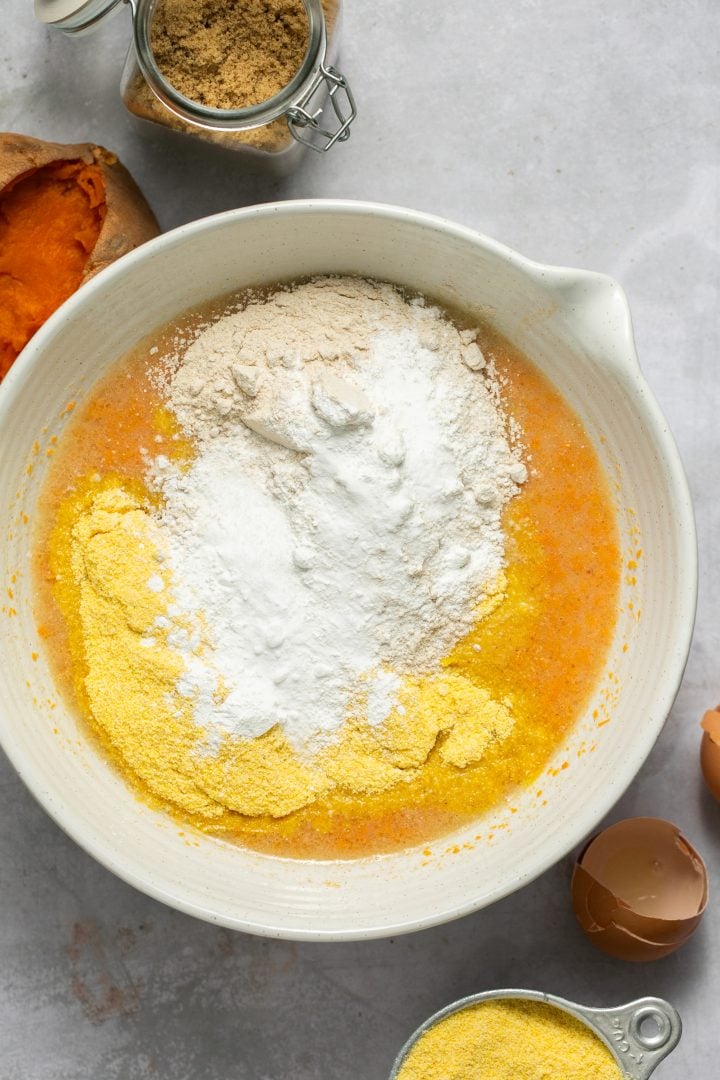 Cornmeal, whole wheat flour, baking powder, baking soda, and salt on top of the wet ingredients for sweet potato cornbread in a white mixing bowl.