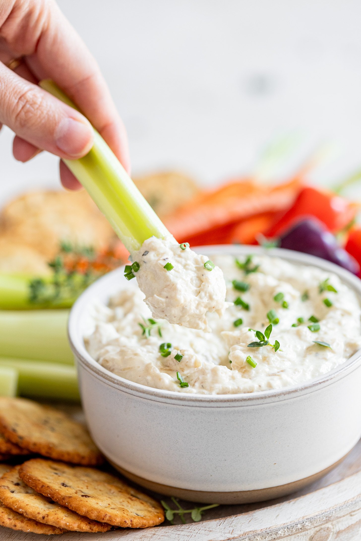 A hand holding a piece of celery being dipped into a white bowl of caramelized onion dip. The bowl is sitting on a plate on a table and is surrounded by out-of-focus red and purple peppers, celery, sprigs of thyme, and crackers. 