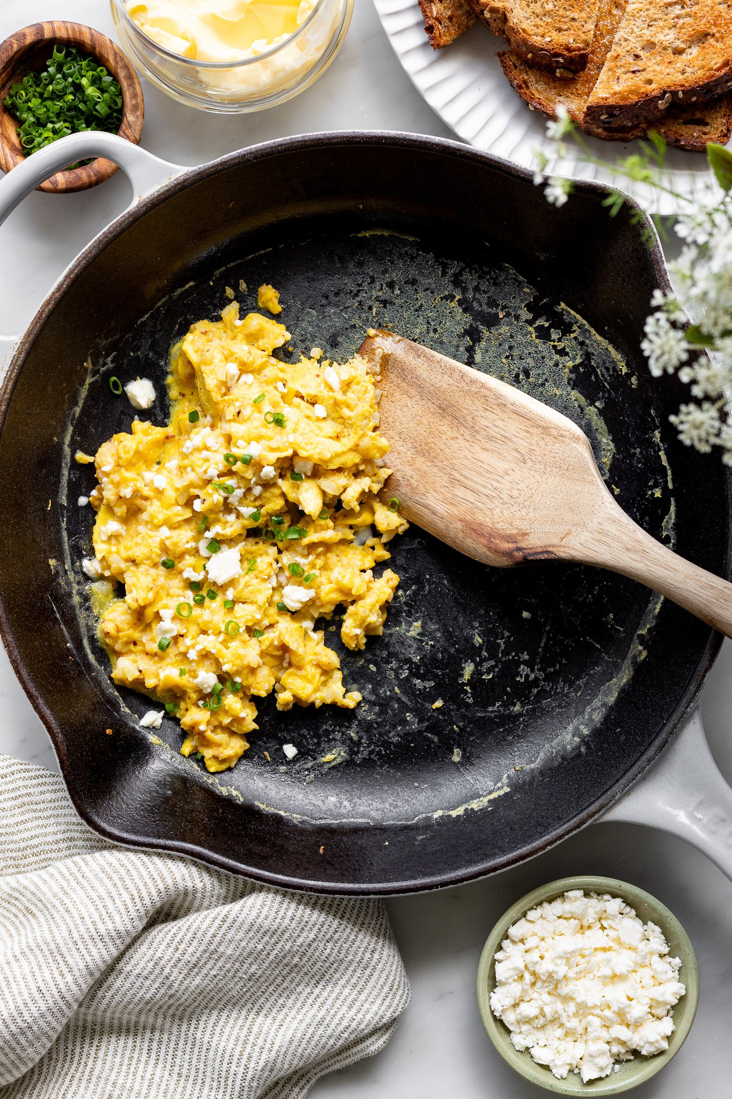 A wooden spatula is stirring cooked scrambled eggs with cauliflower rice in a cast iron pan. There is crumbled feta and chopped chives on top of the eggs. The pan is sitting on a table. There is a napkin next to the pan on the table. There are small bowls of crumbled feta, chopped chives, a glass container with butter, a plate with toast, and a vase with flowers slightly surrounding the plate, slighlty out of frame.
