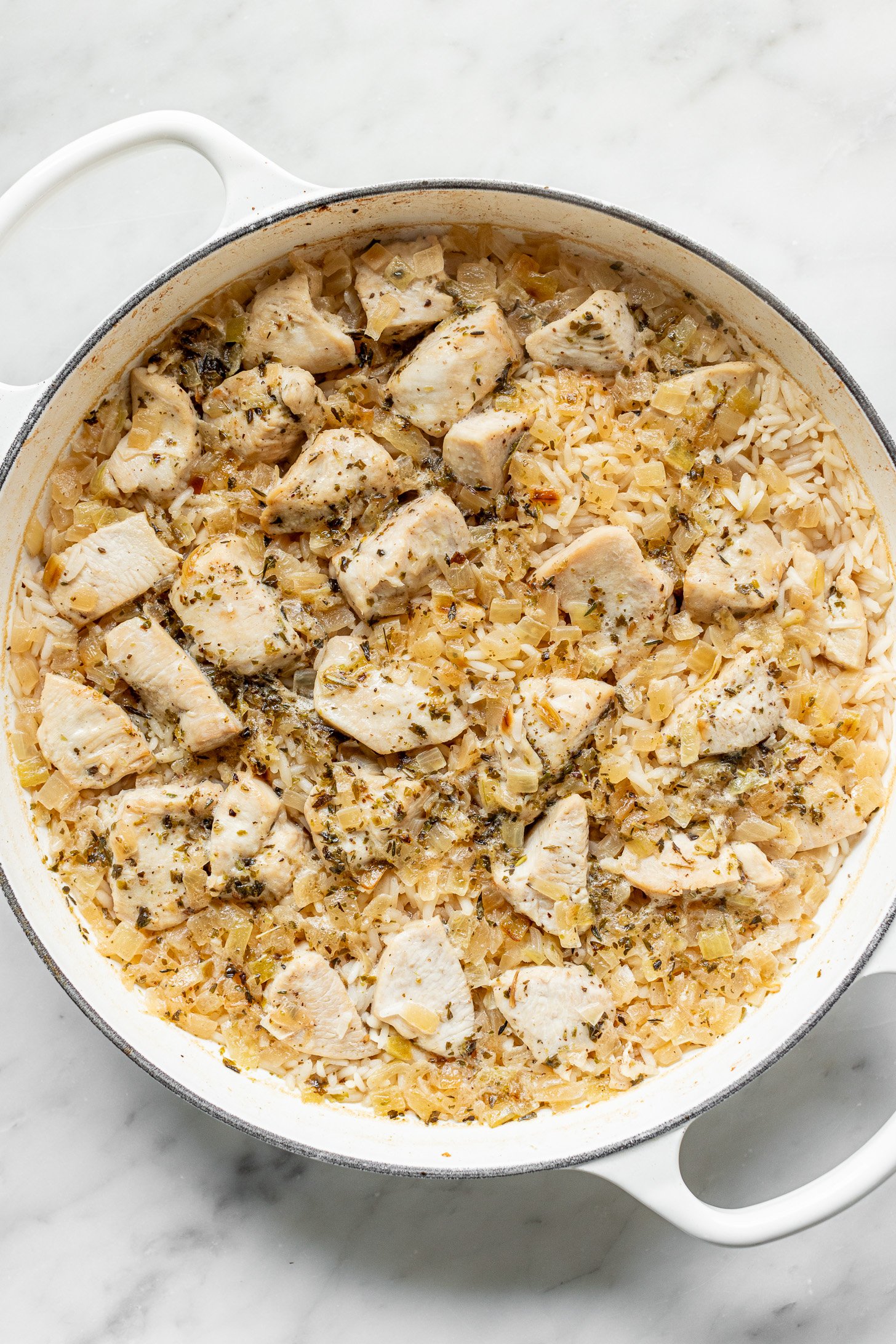 Chicken broth, rice, cooked chicken breast chunks, sauteed onions, and seasonings all mixed together and cooked so liquid has been absorbed in a white dutch oven. Dutch oven is sitting on a white countertop.
