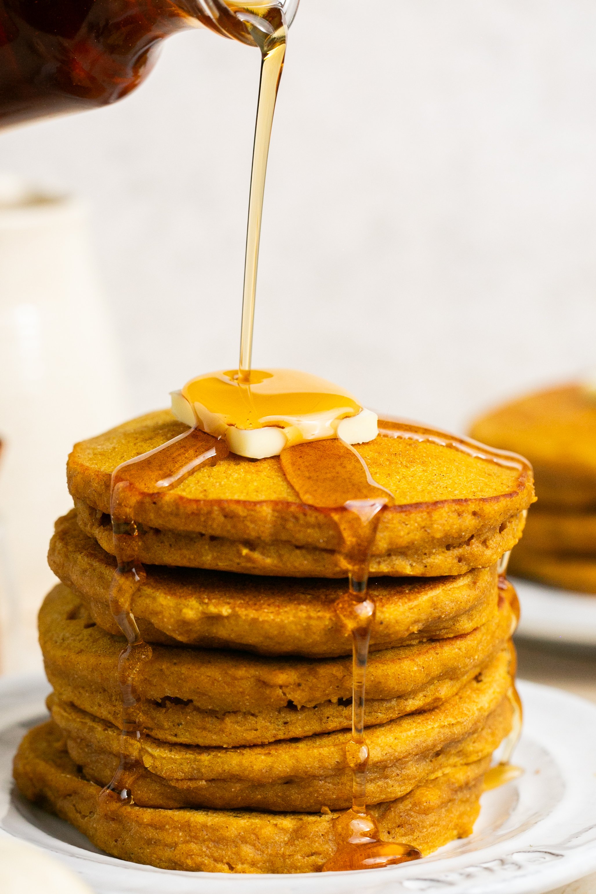 A stack of pumpkin pancakes on a plate with a pat of butter. Maple syrup is being poured over the stack.