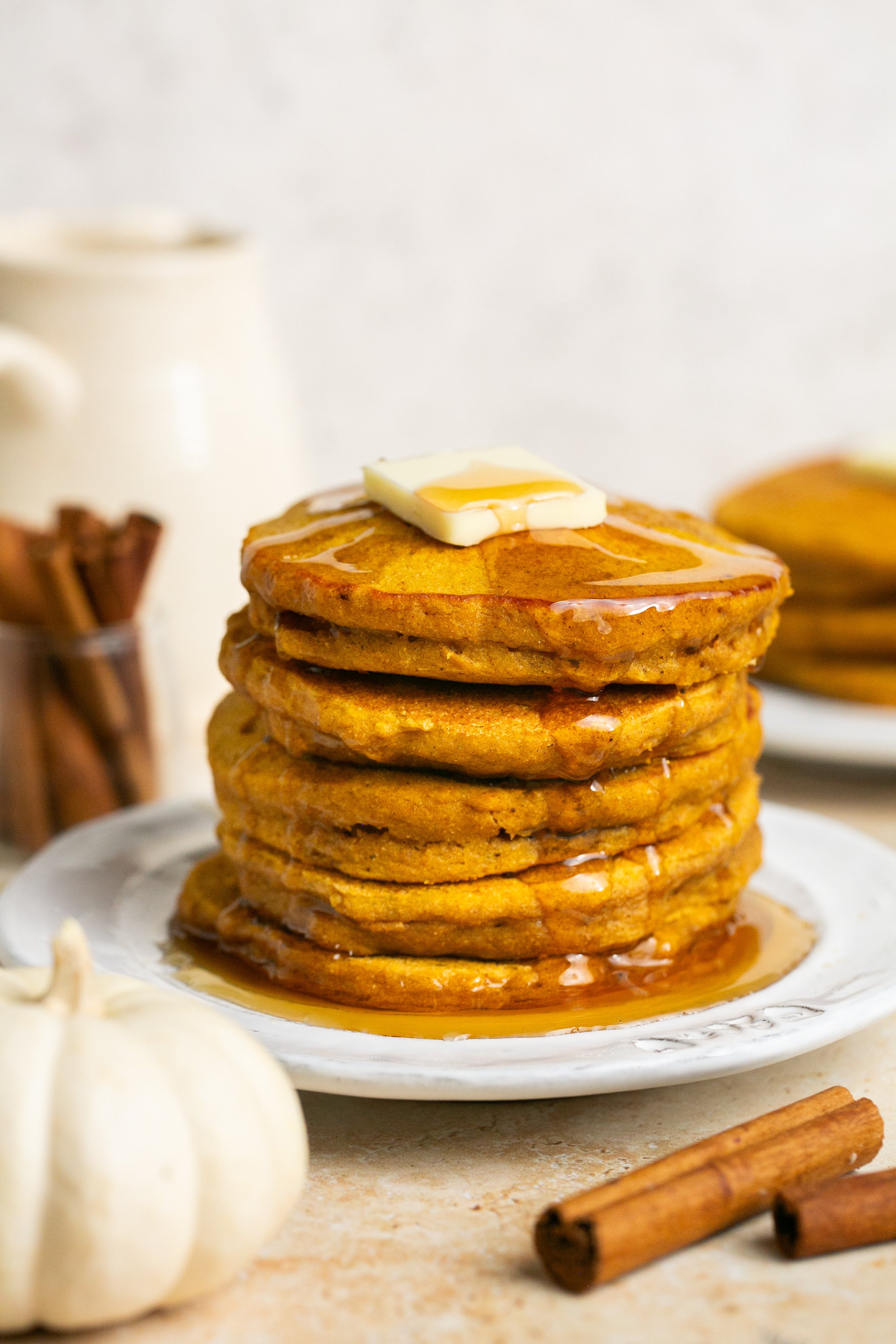 A stack of healthy pumpkin pancakes on a plate topped with a slab of butter and drizzled with maple syrup. There are cinnamon sticks, a white pumpkin, and another stack of pancakes blurred around the main stack.