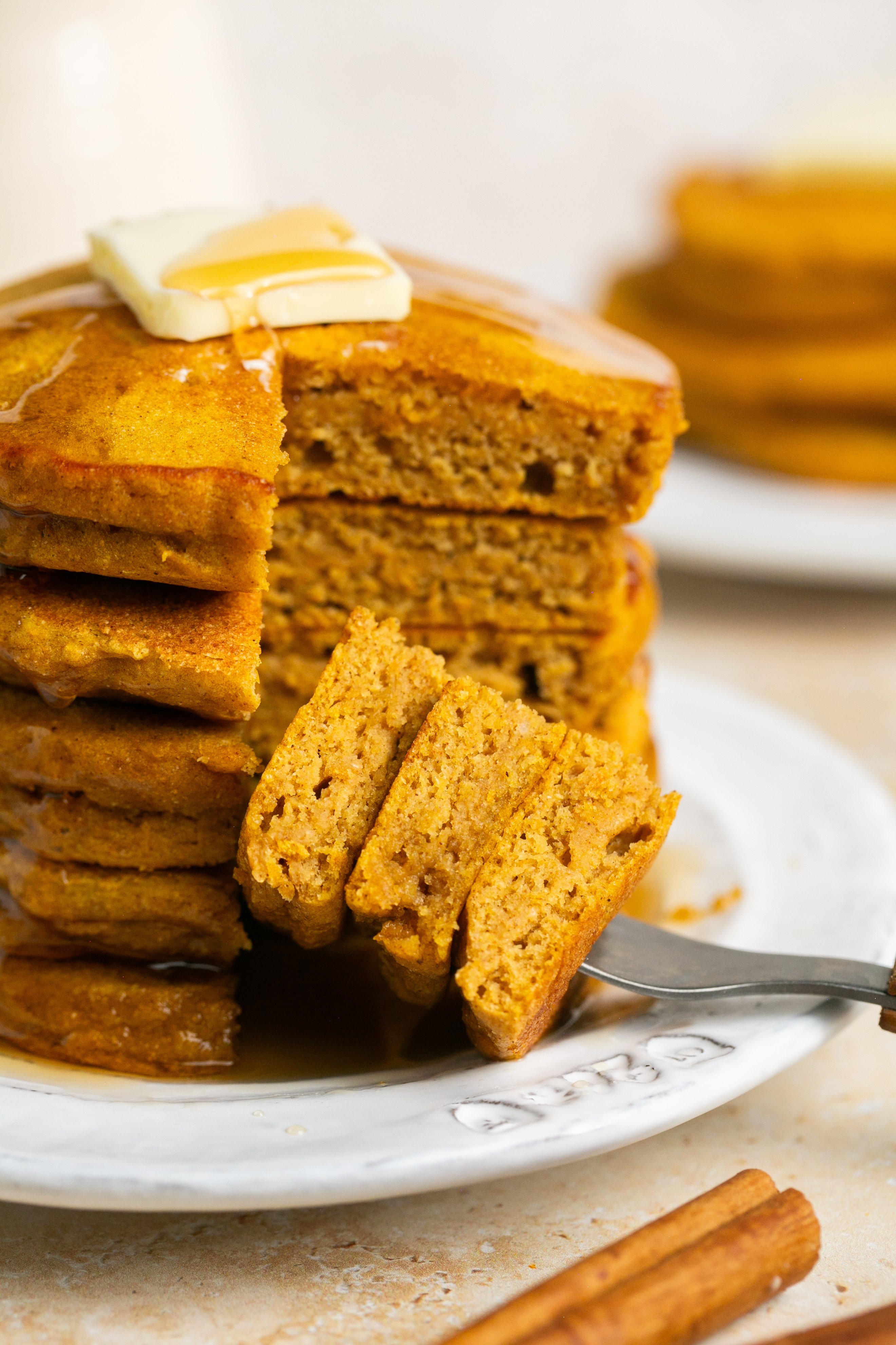 A stack of pumpkin pancakes with a slice taken out of them and on a fork on the plate. The stack is topped with a pat of butter and is drizzled with maple syrup.