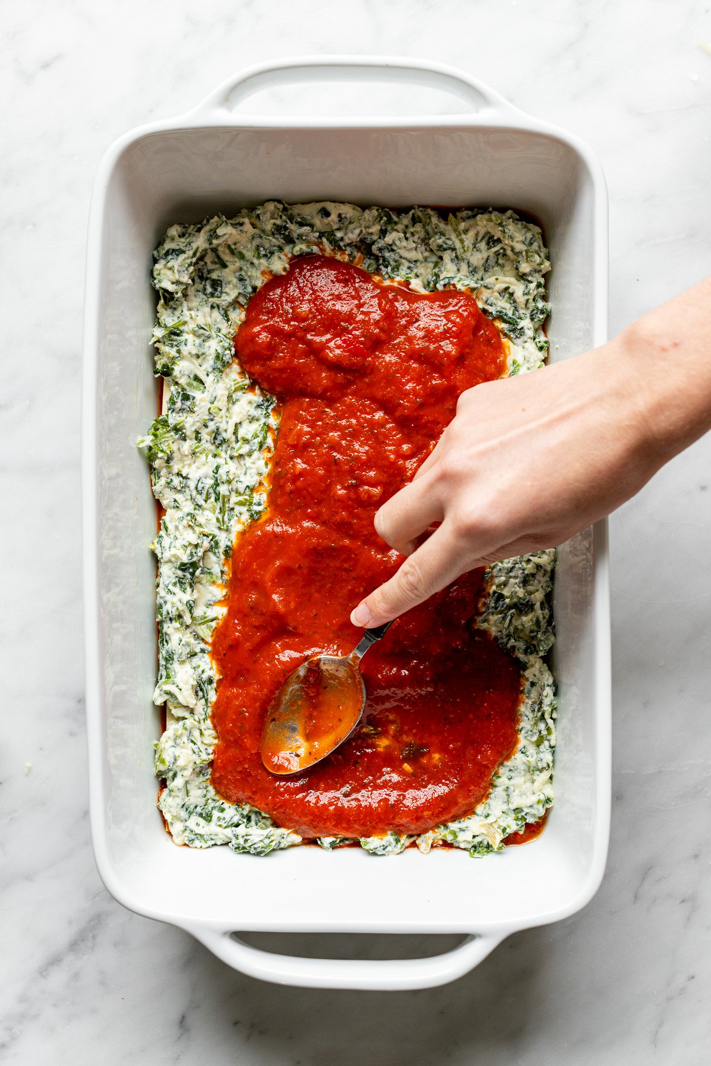 A hand holding a spoon is spreading marinara sauce over the ricotta mixture in a white baking dish.