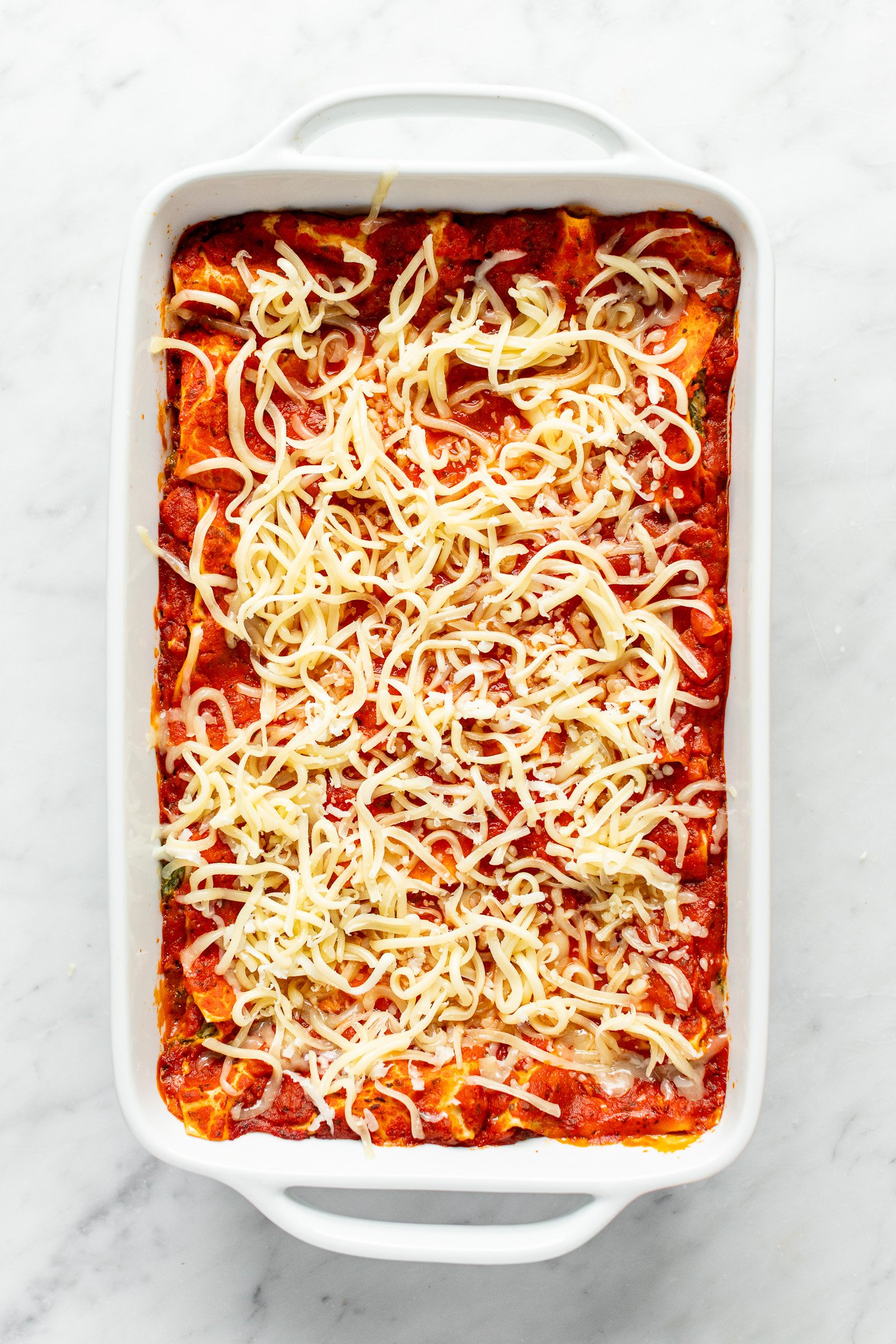Shredded mozzarella cheese is sitting on top of cooked lasagna in a white baking dish.