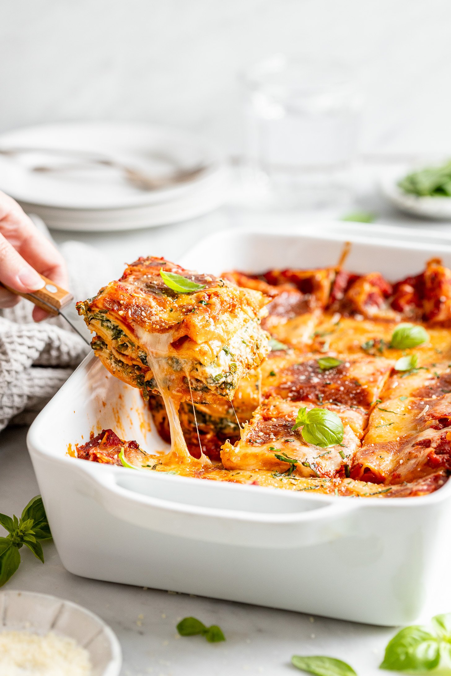Hand holding a serving utensil taking a piece of lasagna out of a white casserole dish. Plates, utensils, basil, a napkin, and a small bowl of parmesan cheese all surrounding lasagna and sitting on a white counter