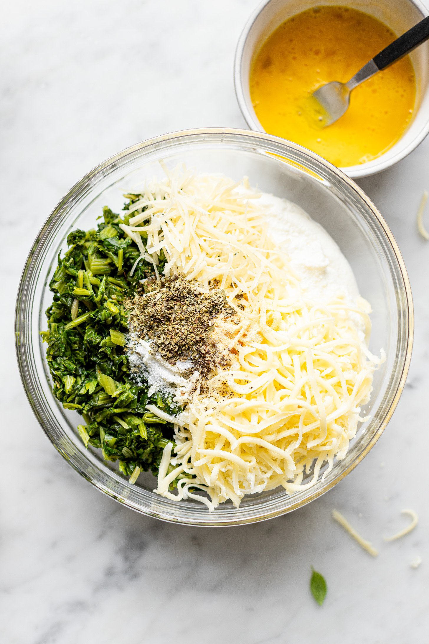 A glass bowl filled with spinach, ricotta, mozzarella cheese, parmesan, garlic powder, Italian seasoning, salt and pepper. There is also a white bowl filled with beaten egg and a fork in the bowl. Both bowls are sitting on a white counter.