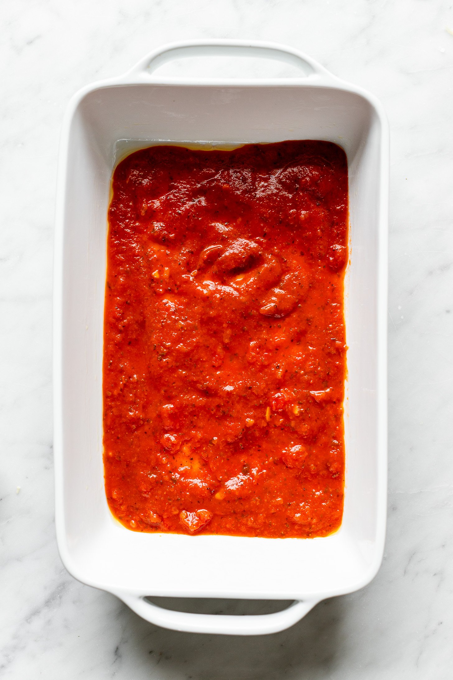 A white baking dish covered with a layer of marinara sauce at the bottom. 